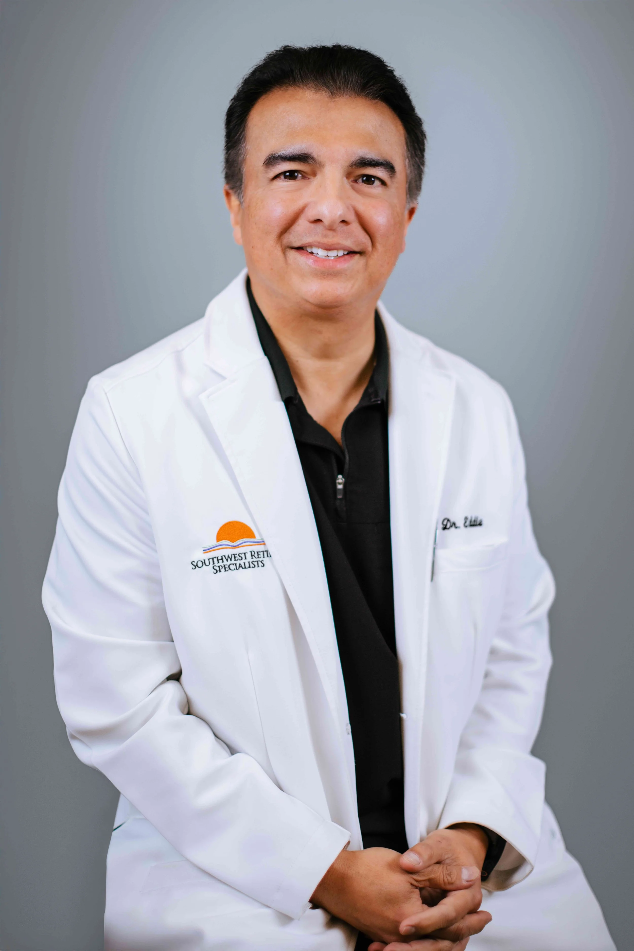 Portrait of a smiling man in a white medical coat with "Southwest Retina Specialists" embroidered on it, standing against a gray background.
