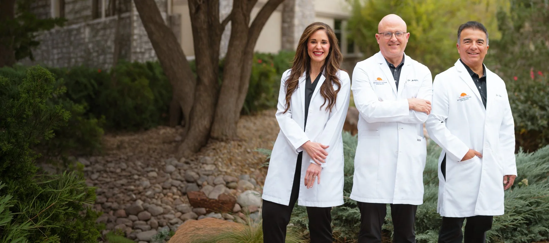 Three healthcare professionals wearing white coats standing outside among trees and plants, smiling at the camera.
