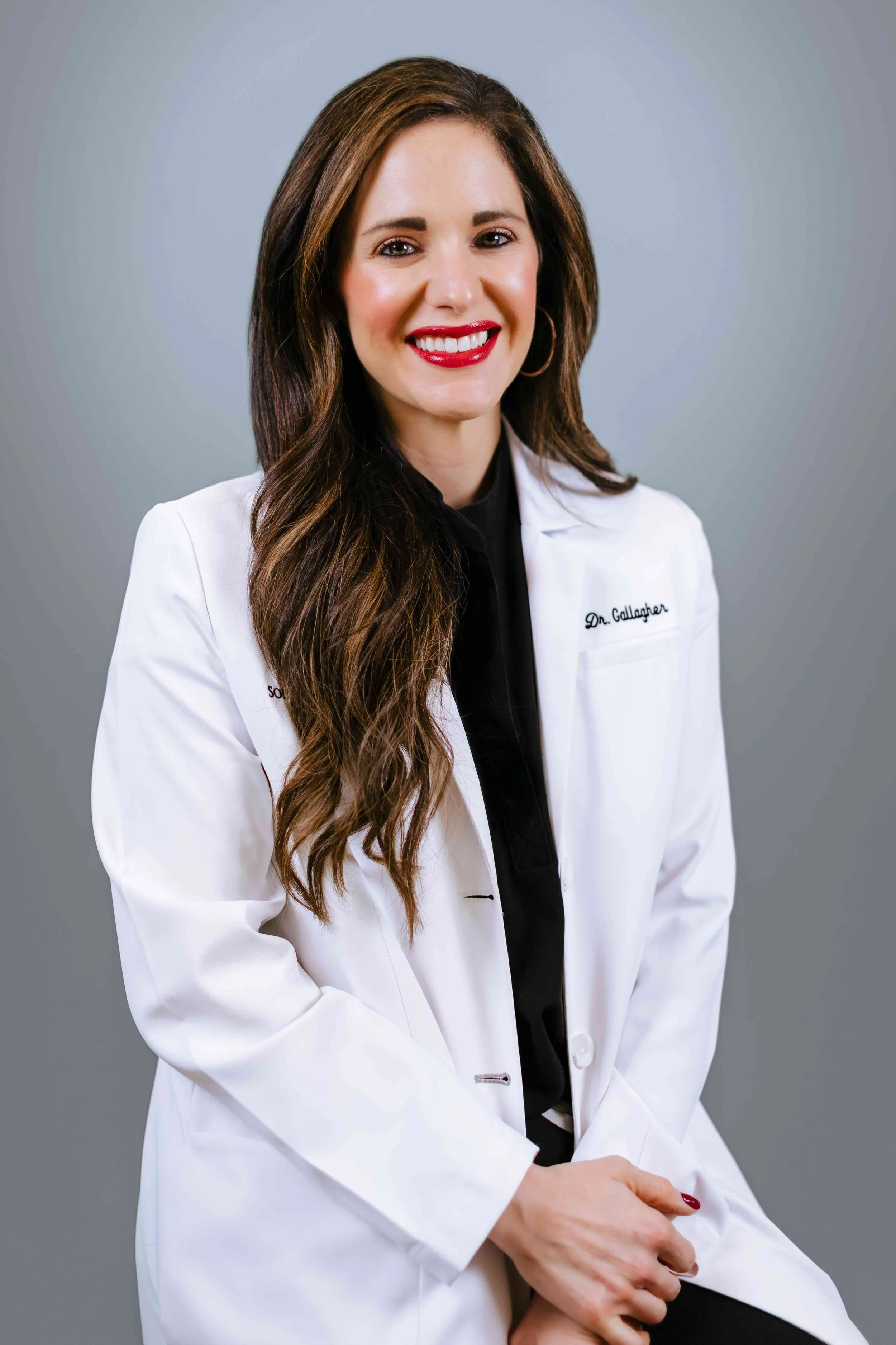 Portrait of a young woman with long brown hair in a white lab coat with "Southwest Retina Specialists" embroidered on it, seated against a gray background.