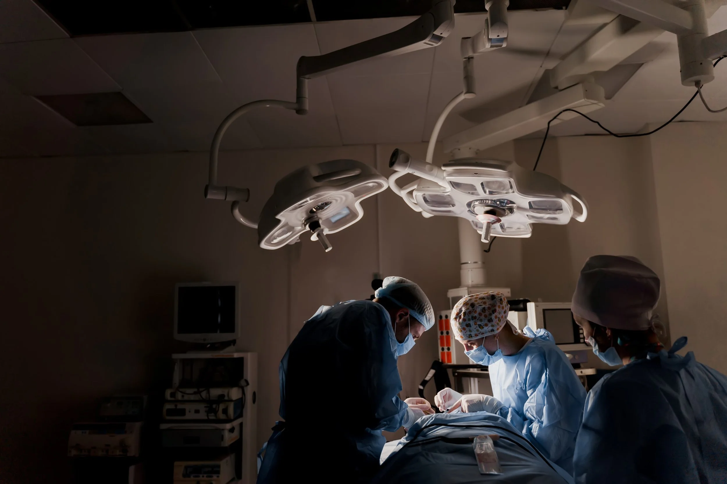 Medical team performing surgery in an operating room with surgical lights overhead and monitors in the background.