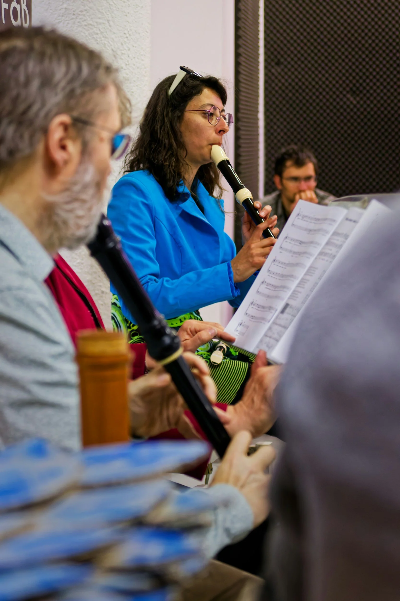 A group of people, including a woman with glasses playing a recorder, practicing music together in a room.