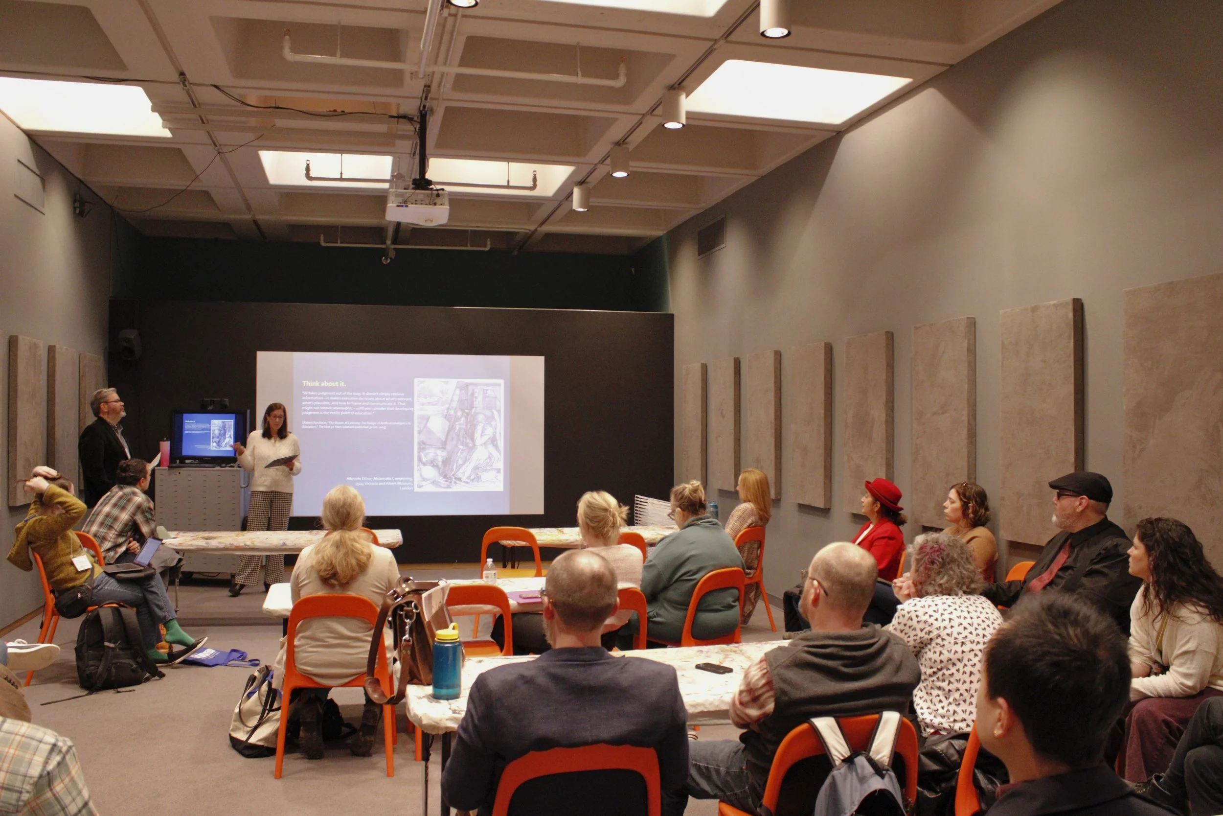 A group of people attending a presentation in a room with a presentation slide projected on the wall. The slide has text and images. Two presenters, a man and a woman, are standing in front of the room, speaking to the audience.