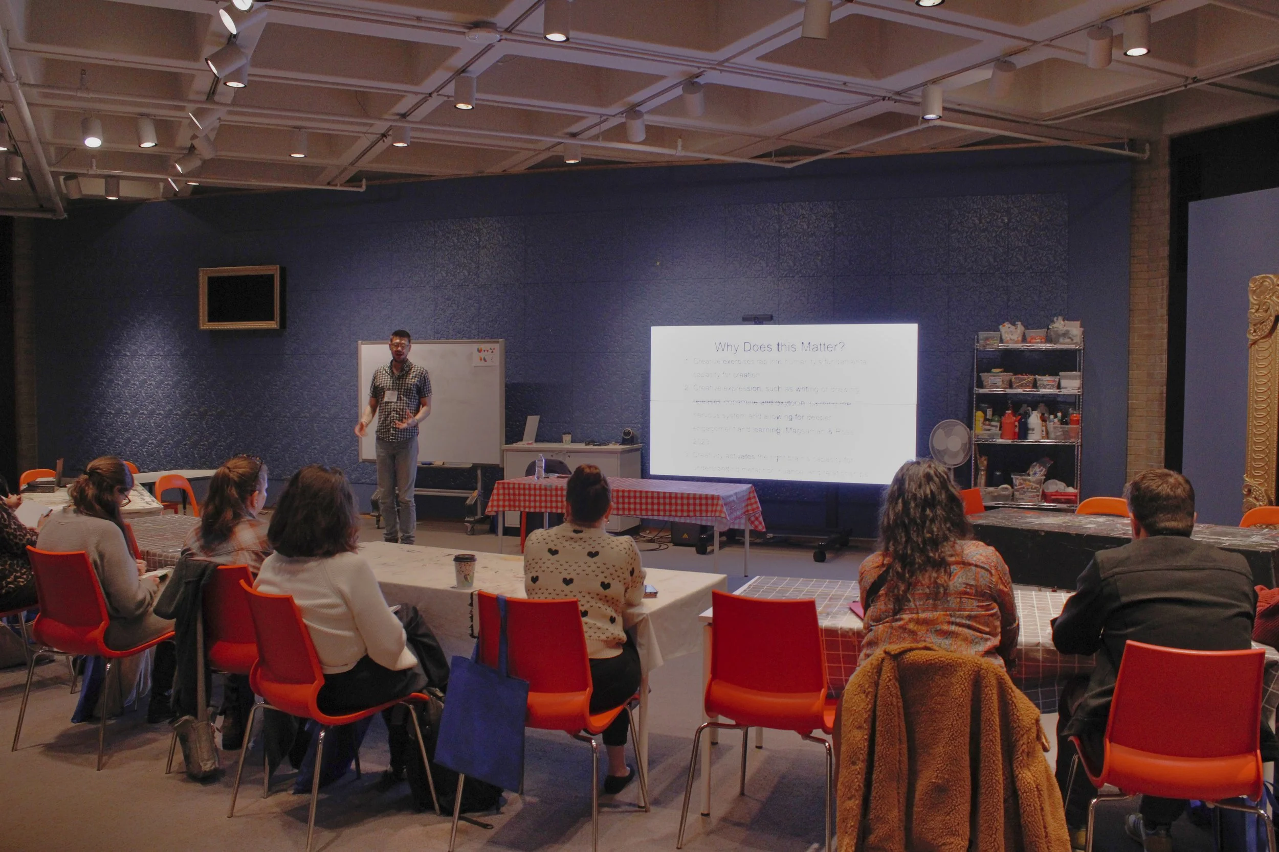 A man standing in front of a whiteboard and a presentation slide in a room with people seated at tables, attending a seminar or workshop.