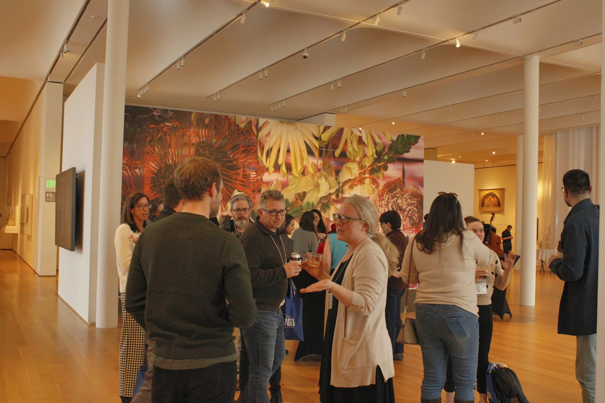 A group of people gathered indoors at an event, engaging in conversation and holding drinks, with a large floral mural on the wall behind them.