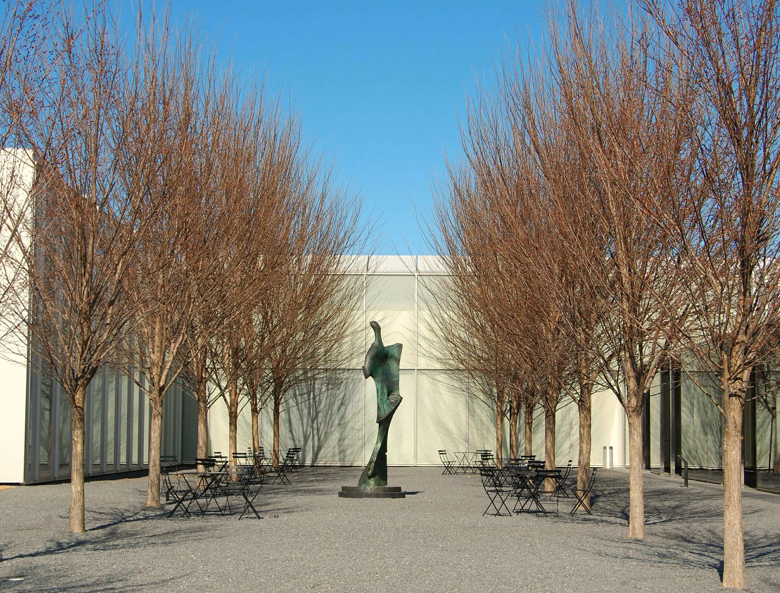 An outdoor public space with leafless trees, metal chairs and tables, a modern sculpture in the center, and a building with glass windows in the background during the daytime.