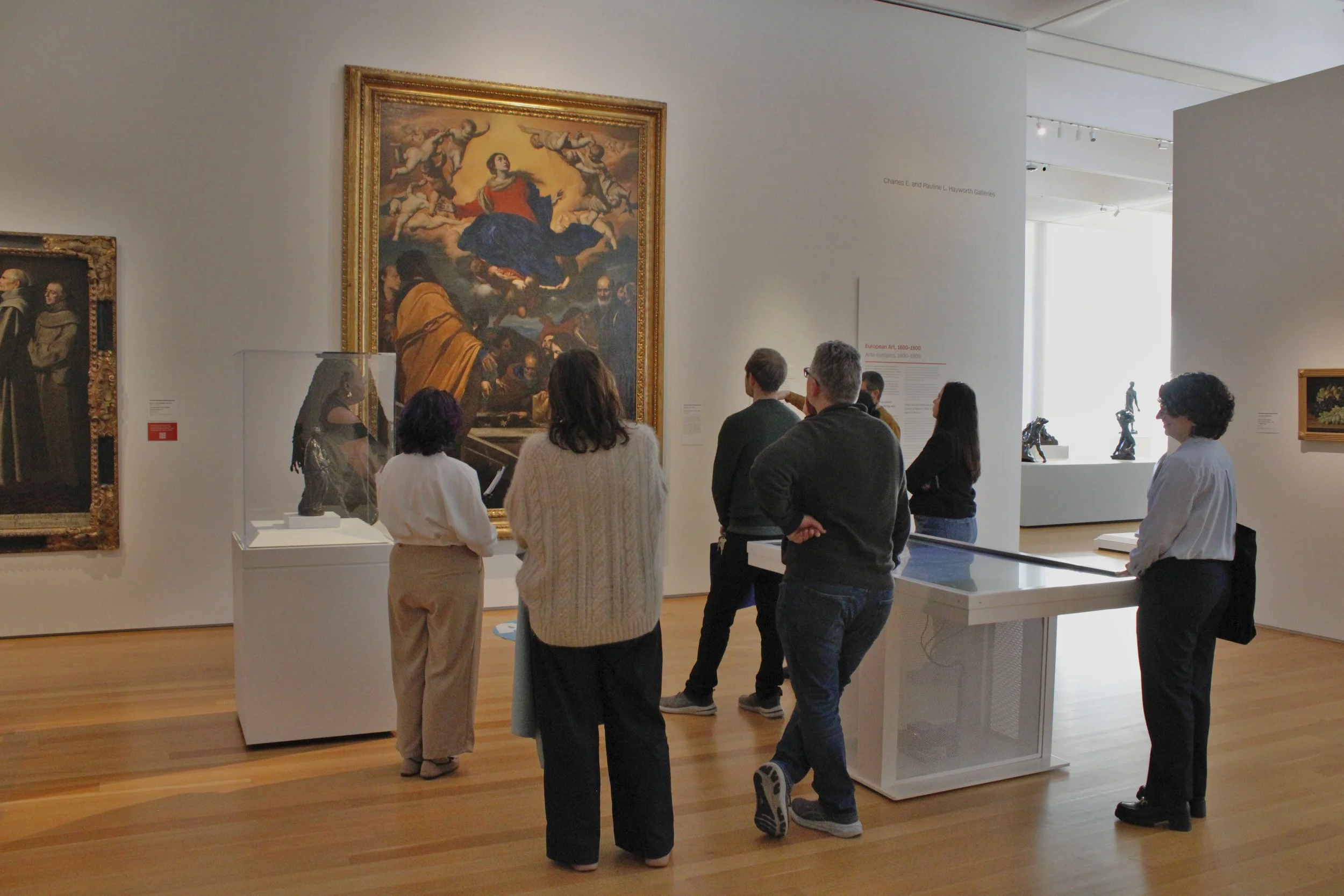 Group of people viewing art exhibit in a gallery, focusing on a large painting of a seated woman with surrounding cherubs.