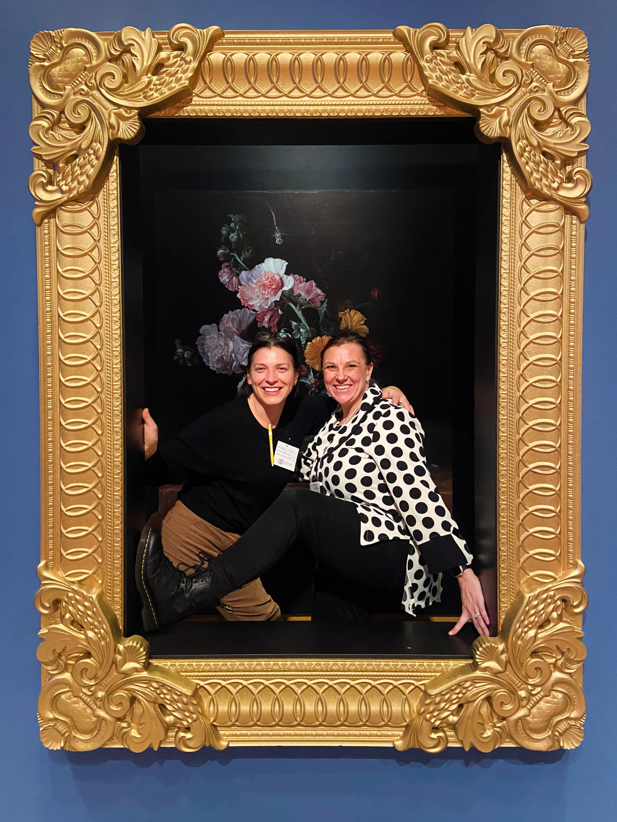 Two women smiling and posing inside a large, ornate gold picture frame with floral artwork background and blue wall.