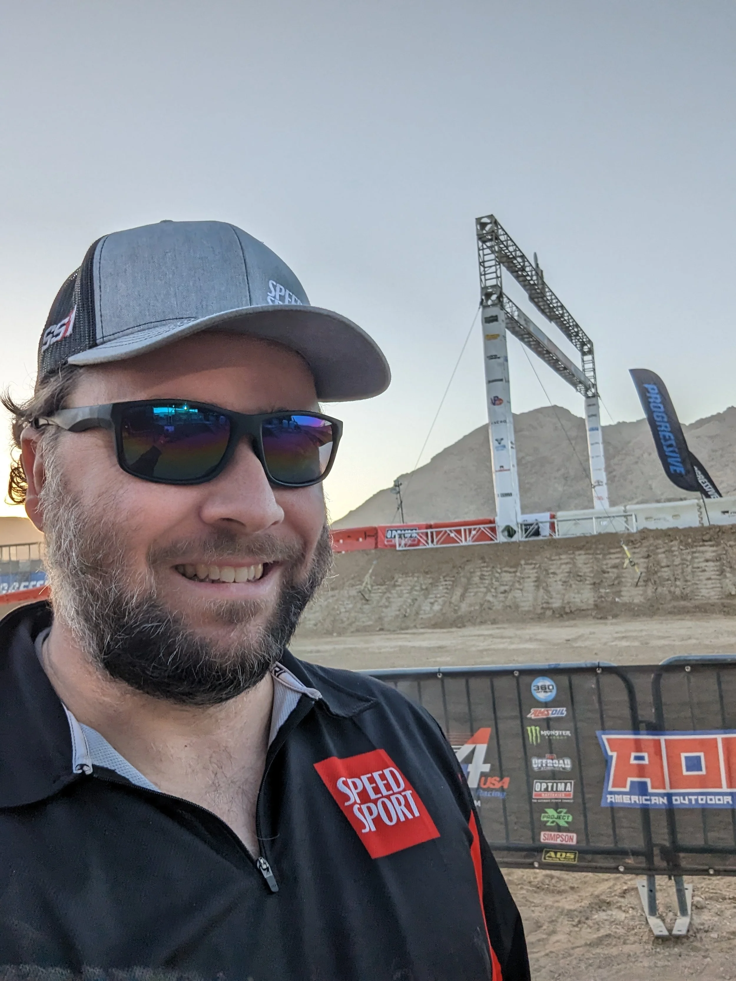 A man, Kurt Shrader, wearing sunglasses, a gray cap, and a black racing jacket with a red 'Speed Sport' logo, smiling at a dirt race track with a mountainous background and a racing event banner.