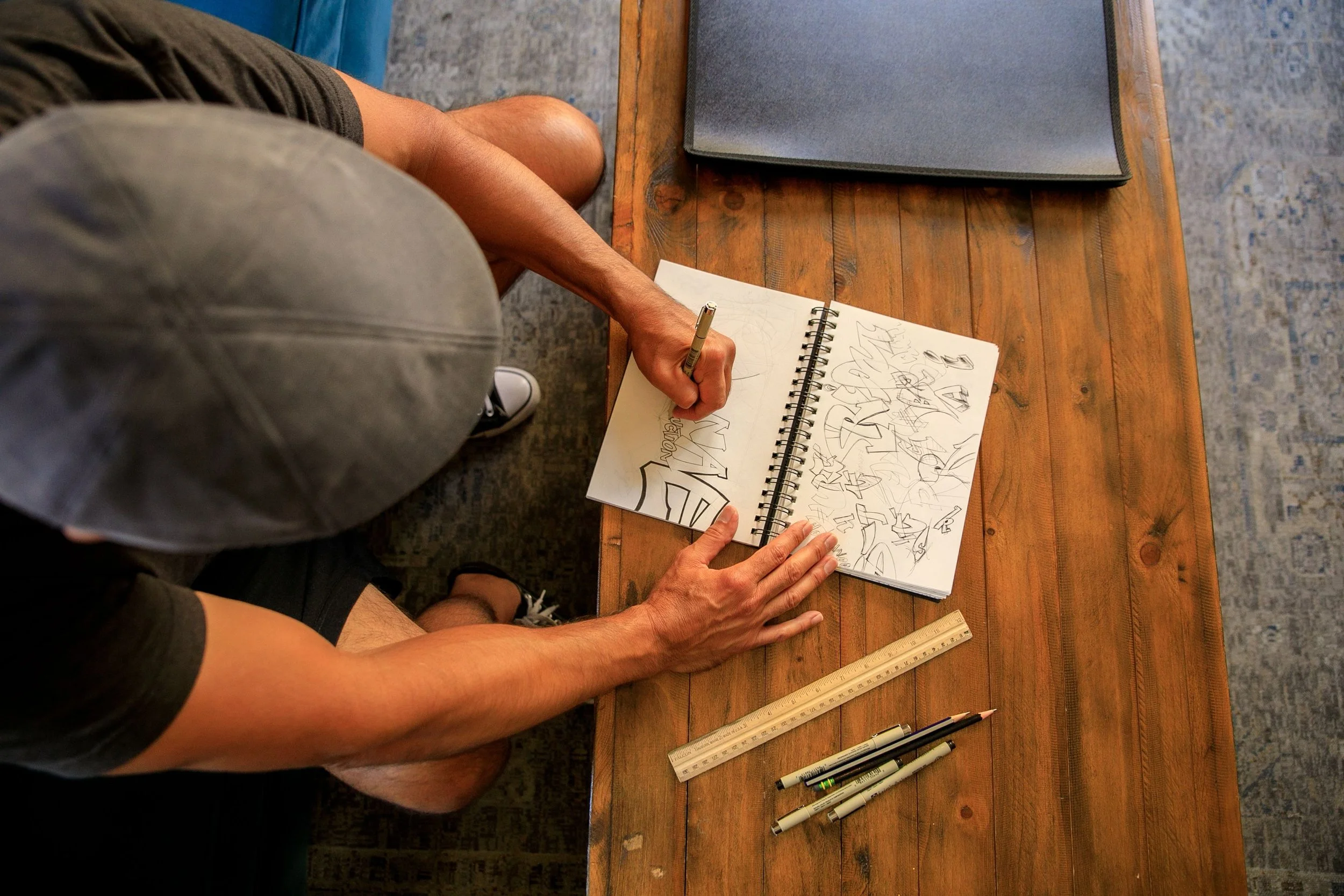 Overhead view of a man drawing in a spiral-bound sketchbook at a wooden table, with pencils and a ruler nearby.