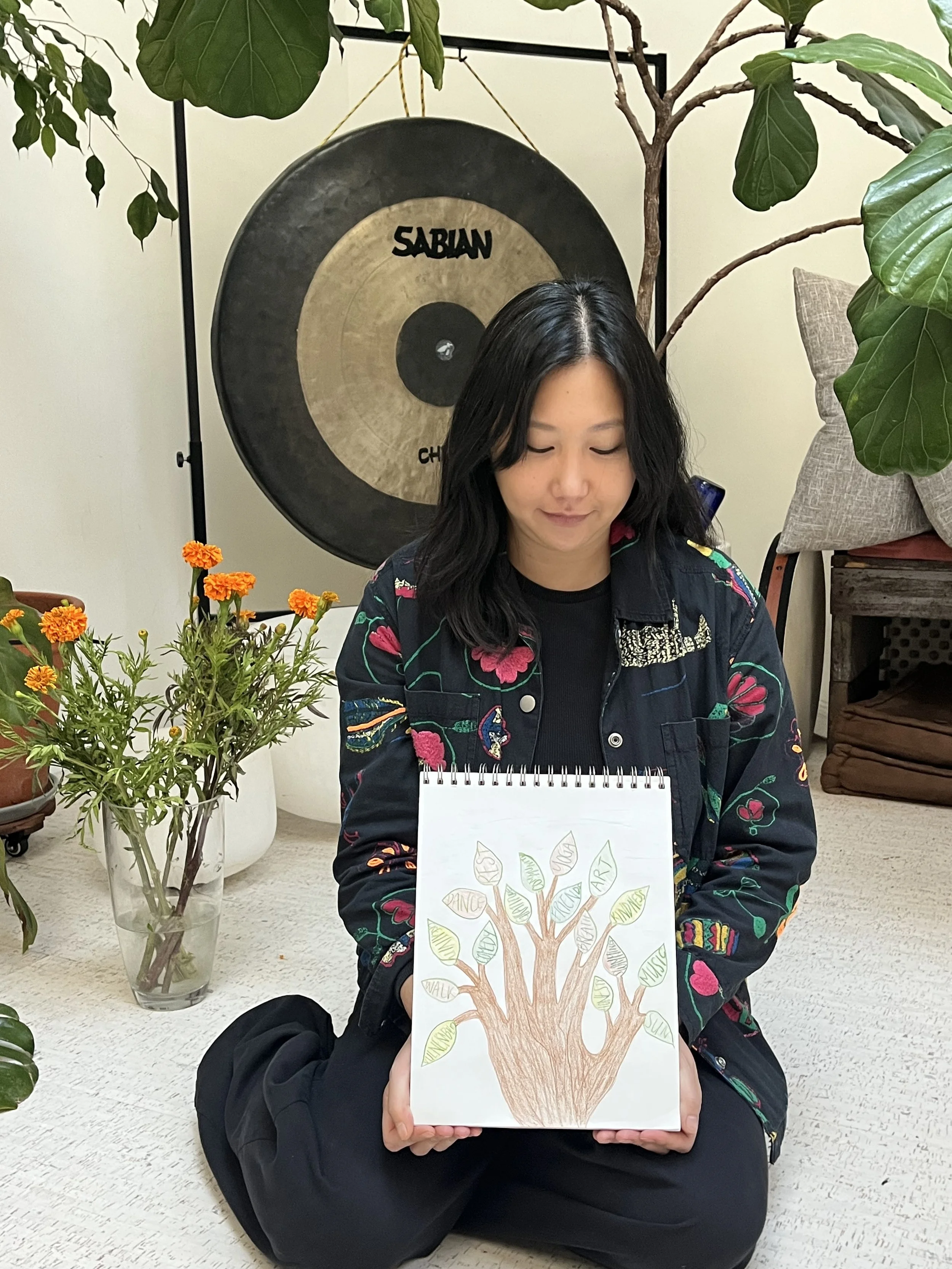 Liz Cho, art therapy intern, seated holding a colorful tree artwork, with leafy plants and a large gong behind her.