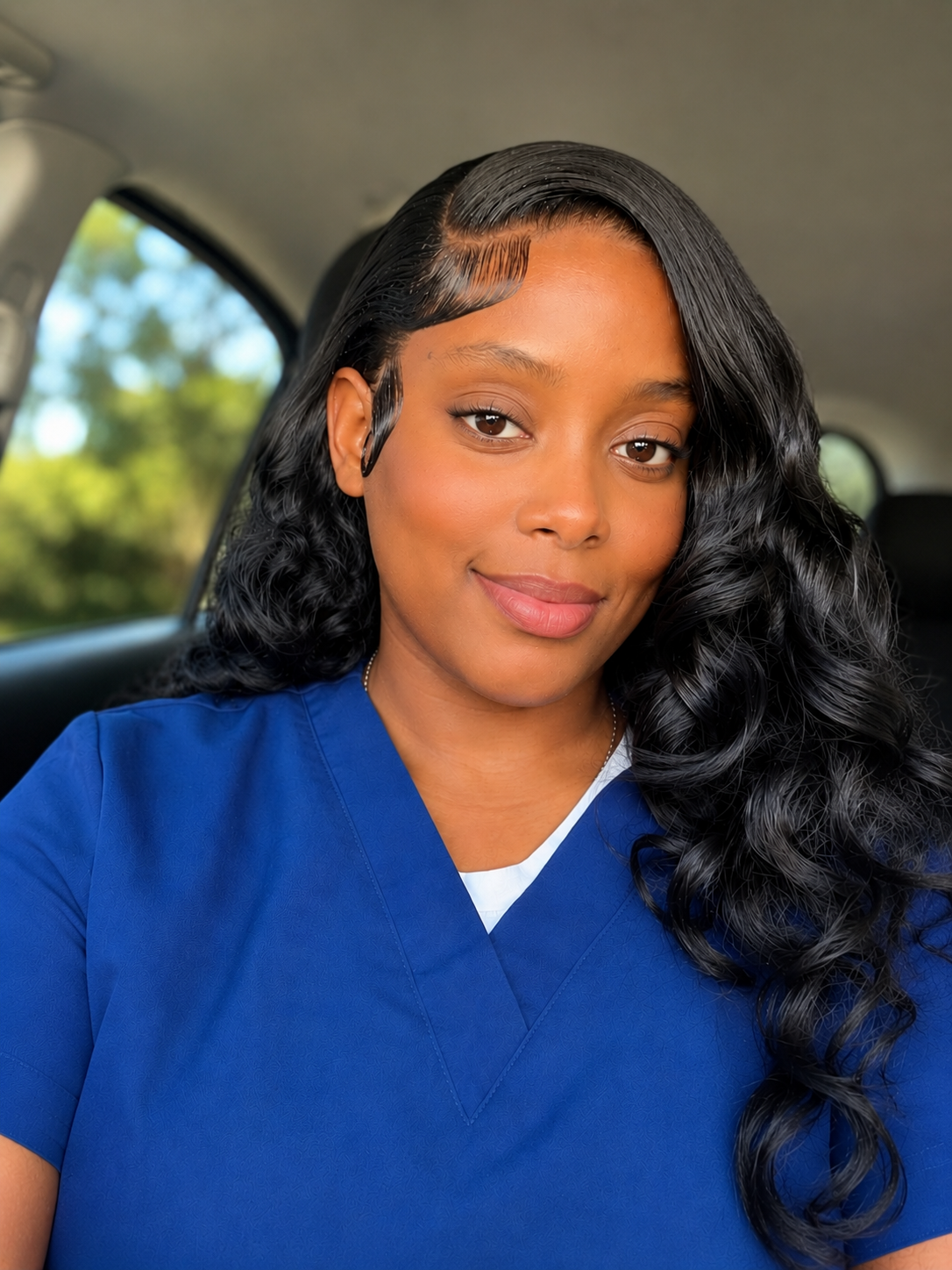 A woman in blue scrubs sitting in a car, smiling at the camera with long black curly hair.