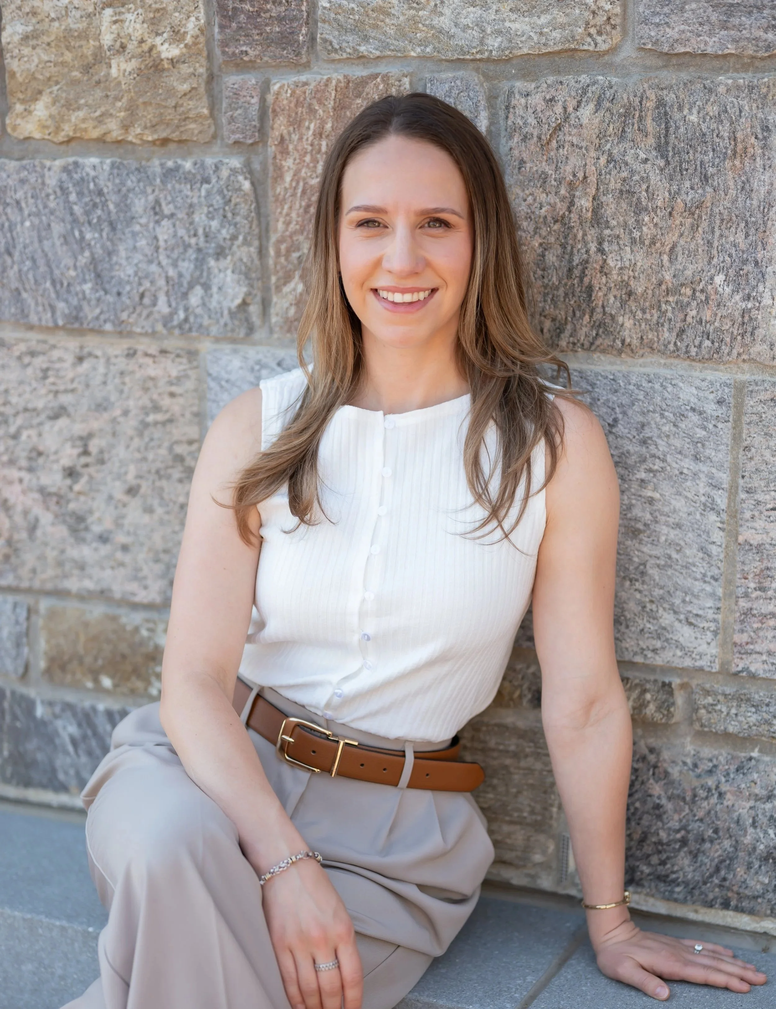 A woman with long brown hair, wearing a sleeveless white top, beige trousers, and brown belt, sitting against a stone wall, smiling at the camera.