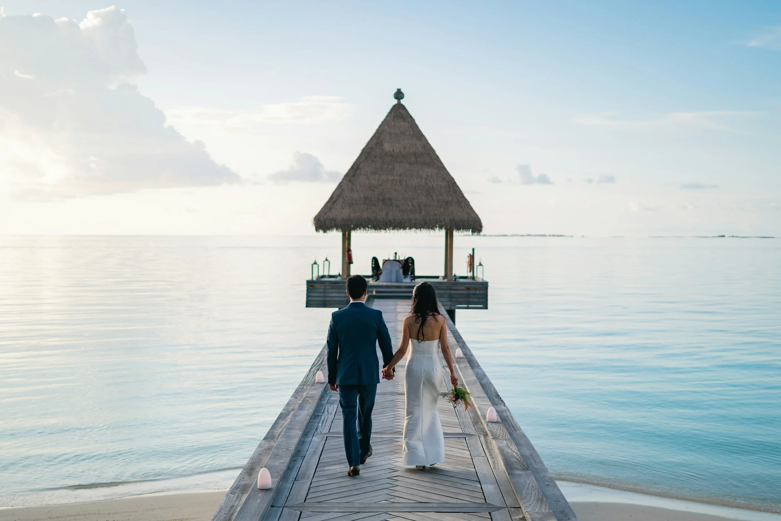 A couple in wedding attire walking hand in hand on a wooden pier towards a thatched-roof gazebo over calm water at sunset or sunrise.