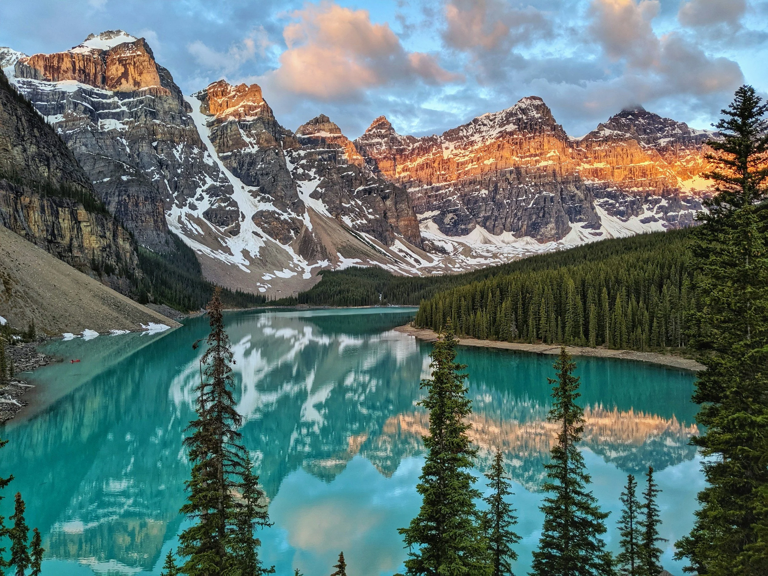 Snow-capped mountains at sunset over a turquoise lake, with a forested shoreline and partly cloudy sky.