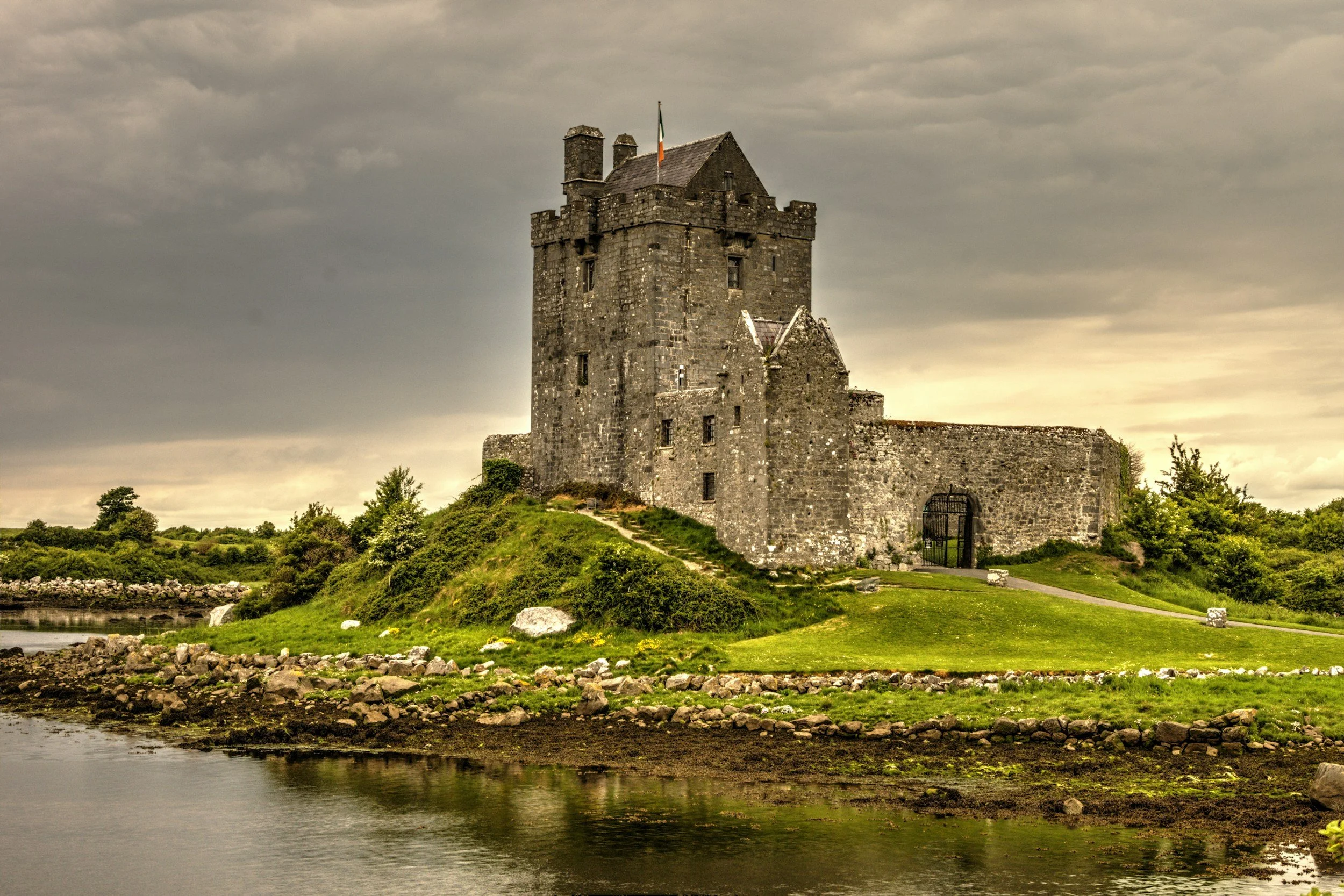A stone castle with a flag on top, surrounded by greenery, beside a river on a cloudy day.