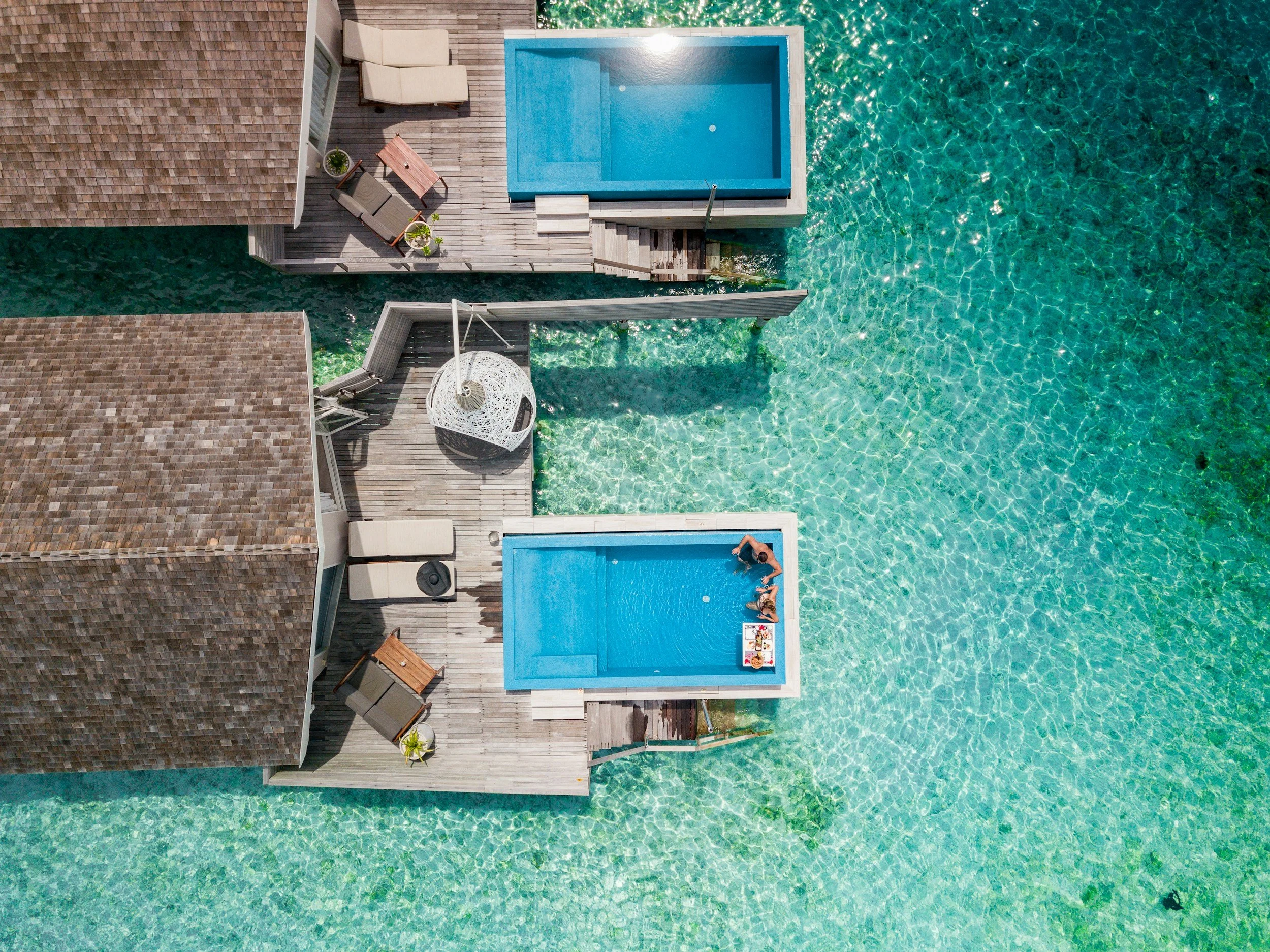 Aerial view of a house with a wooden deck, patio furniture, and two swimming pools, one small and one large, over clear aquamarine water.