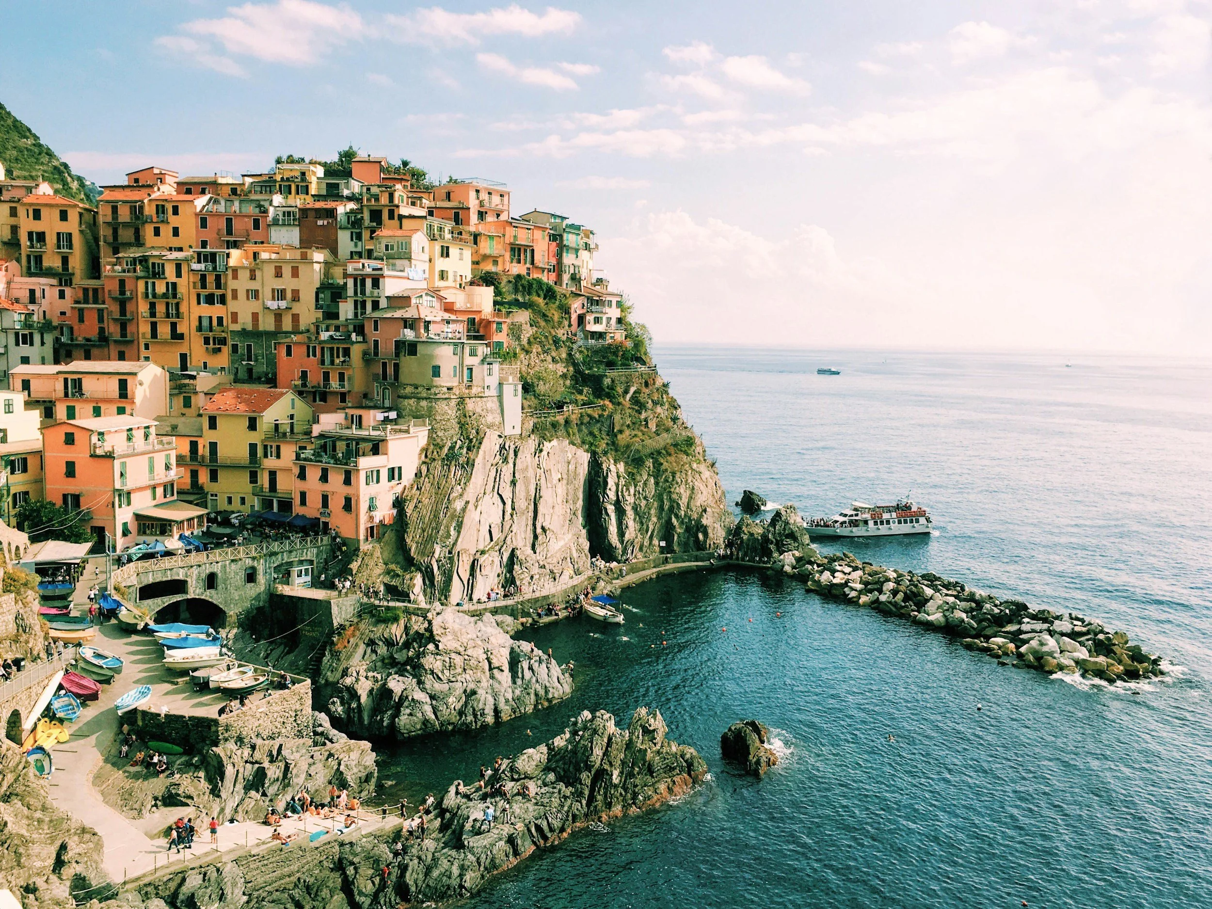 Colorful houses built on a hillside overlooking the sea, with boats docked nearby and a dock with people walking along it.
