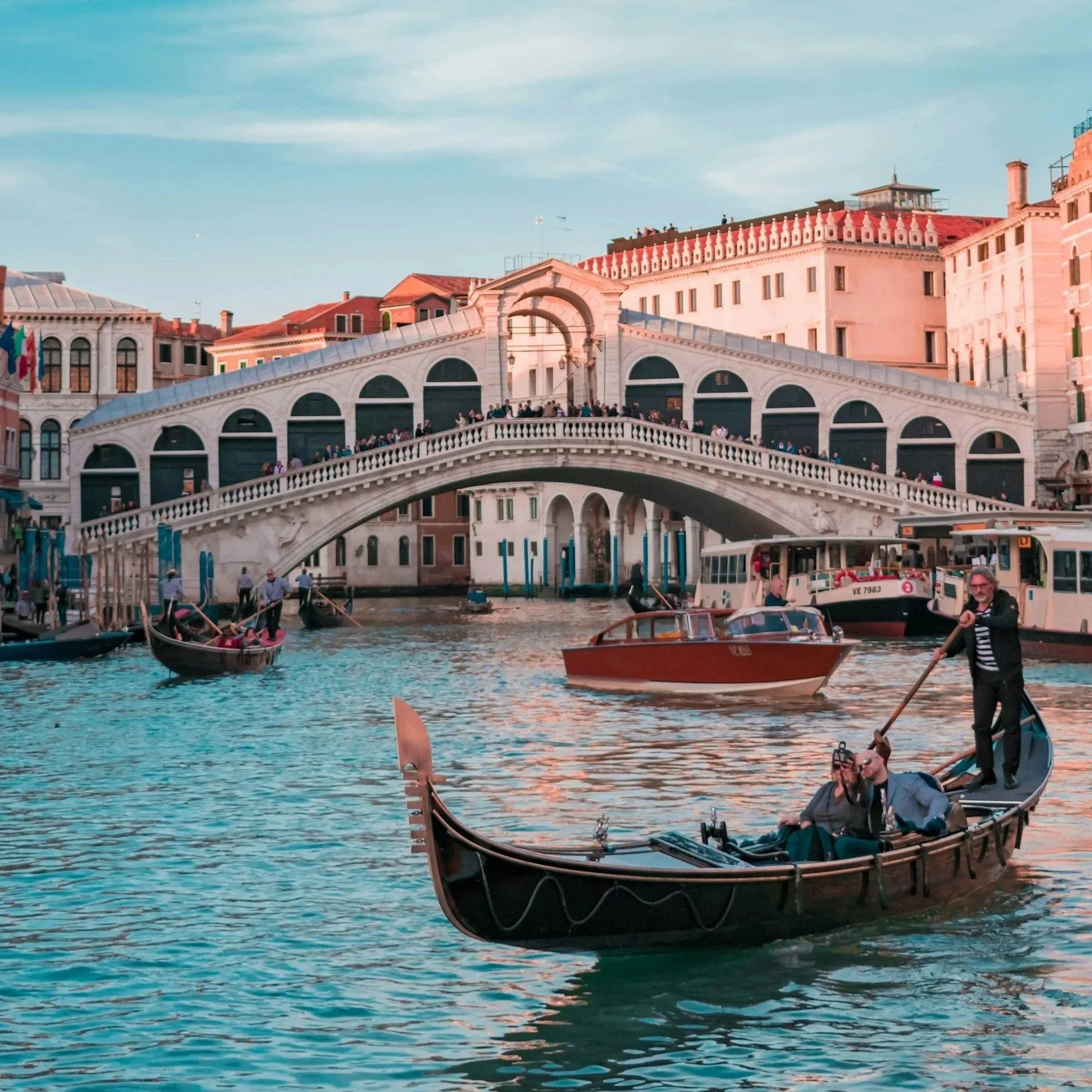 Venetian canal scene with gondolas and water taxis, historic architecture, and a arched bridge with tourists walking across.