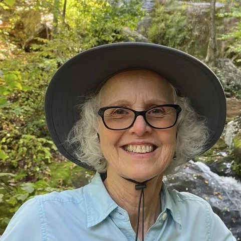 An elderly woman with gray curly hair, wearing glasses, a wide-brimmed hat, and a light blue shirt, smiling outdoors near a stream with rocks and greenery in the background.