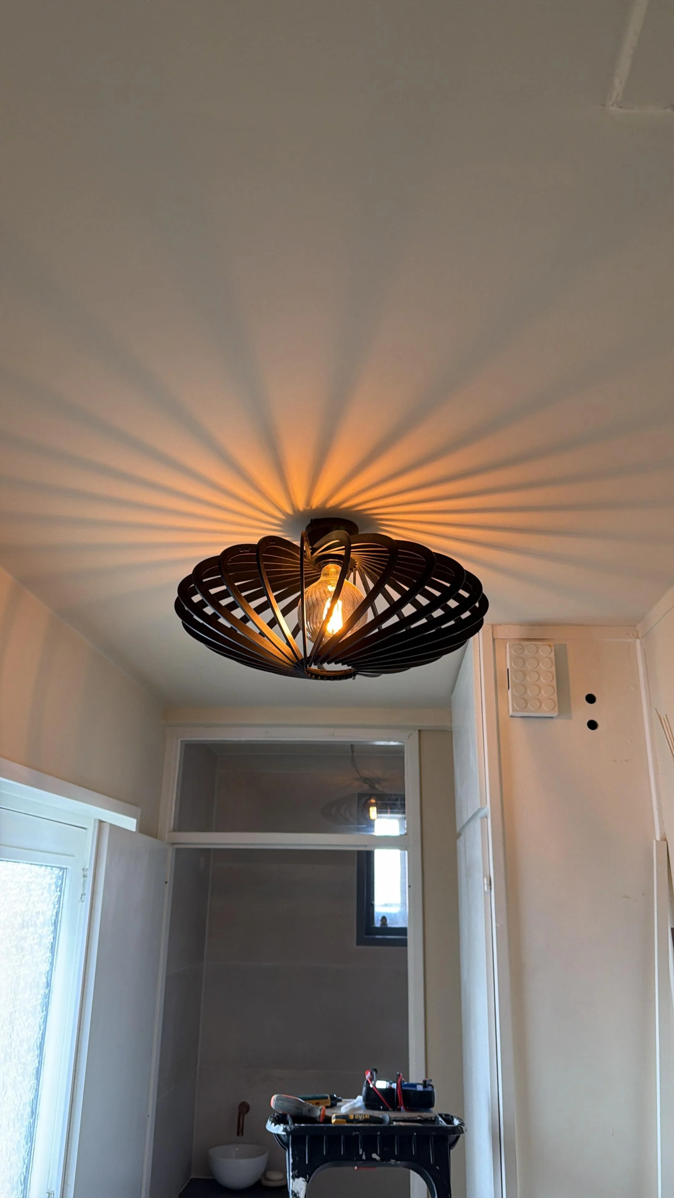 Binnenruimte met een gedraaide plafondlamp die schaduwpatronen op het plafond projecteert, een deur en ramen aan de onderkant, en een tafel met gereedschap.