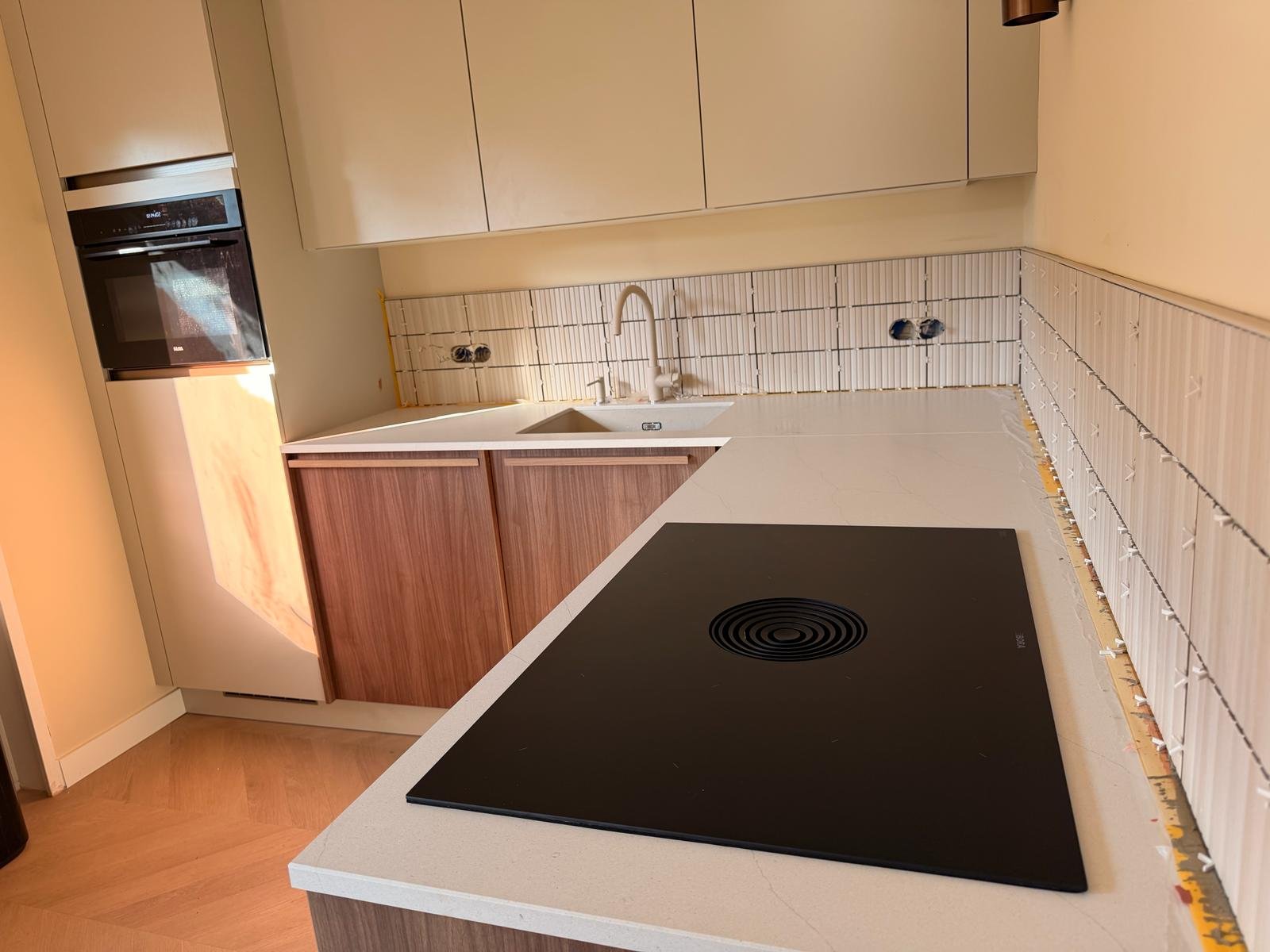 De keuken ondergaat een renovatie met nog ontbrekende achterwandtegels, een fornuis op het aanrecht, een witte spoelbak, houten en wit keukenkastjes, en ingebouwde oven en kookplaat.