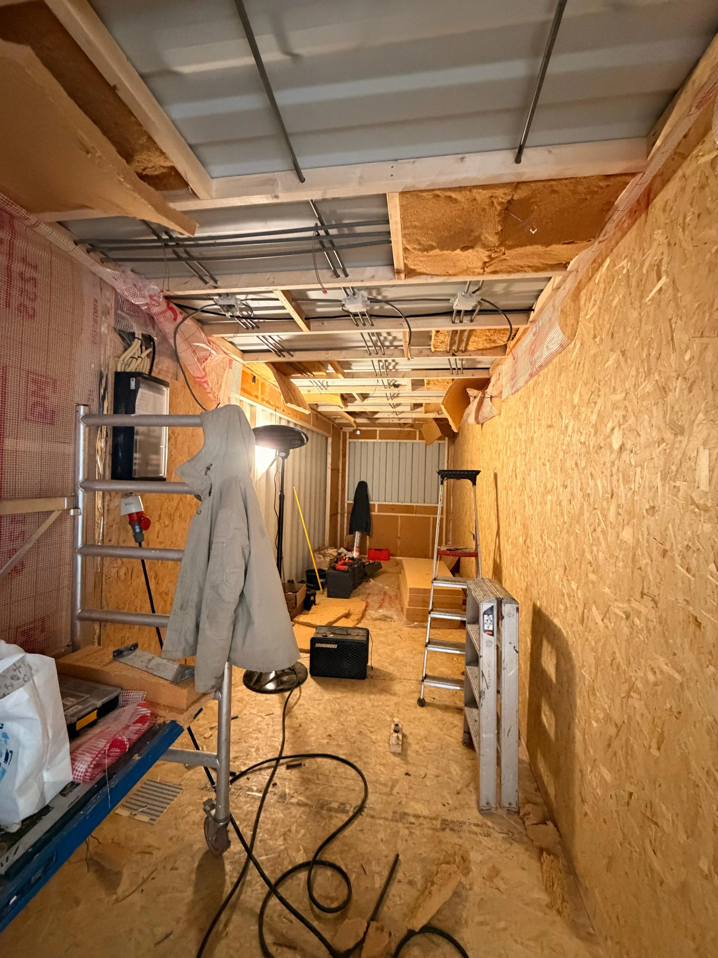 Een kamer onder constructie met houten wanden en isolatiemateriaal. Diverse bouwmaterialen en gereedschap zoals ladders, elektrisch gereedschap, en isolatiemateriaal zijn aanwezig.