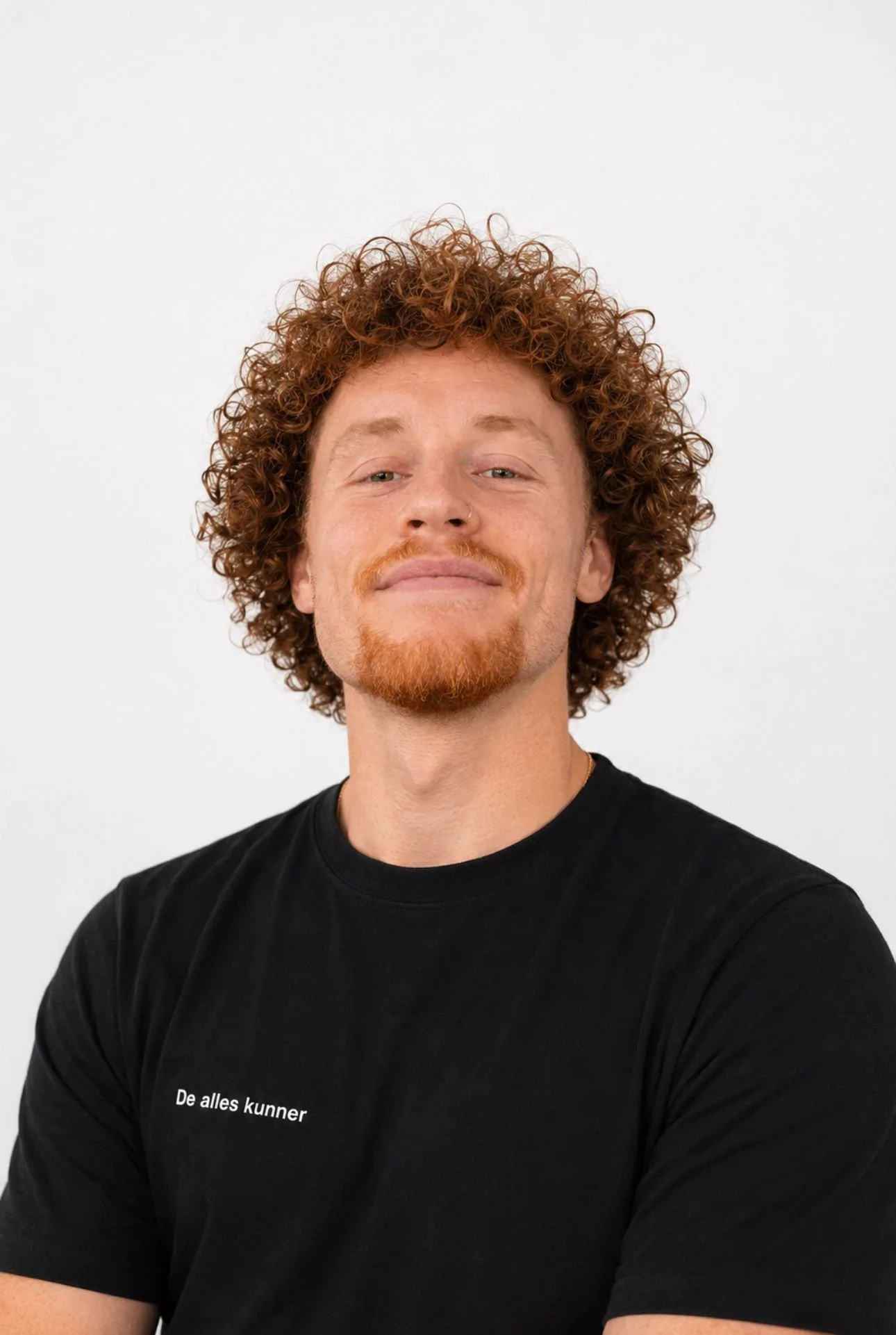 Man met lichtbruine, kroezende haar en een lichte baard, zittend voor een witte achtergrond, draagt een zwarte t-shirt met Nederlandse tekst.