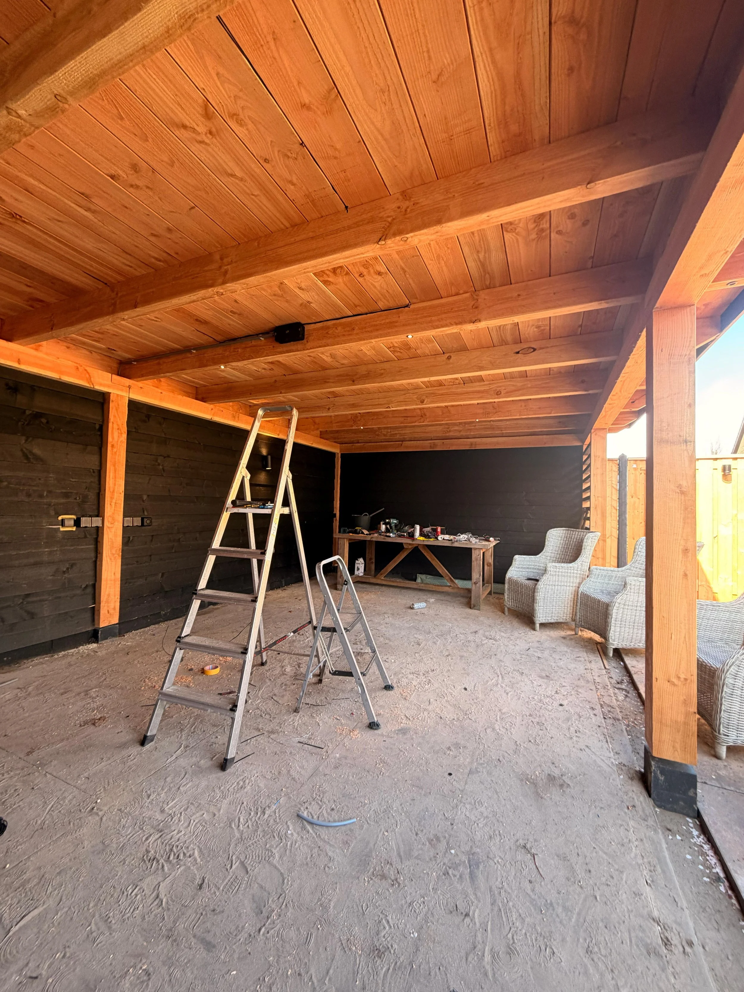 Een overdekte buitenruimte onder constructie met houten plafond en steunbalken. Er staan trappen en er liggen gereedschappen en materialen op de grond en tafels, klaar voor verder werk.