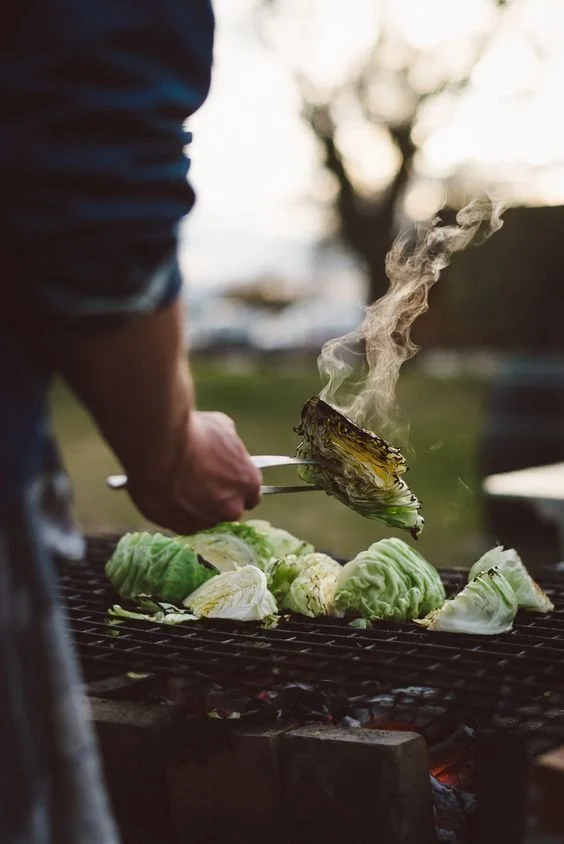 Person grilling lettuce and other vegetables outdoors over an open flame with smoke rising, backlit by a sunset or bright light.