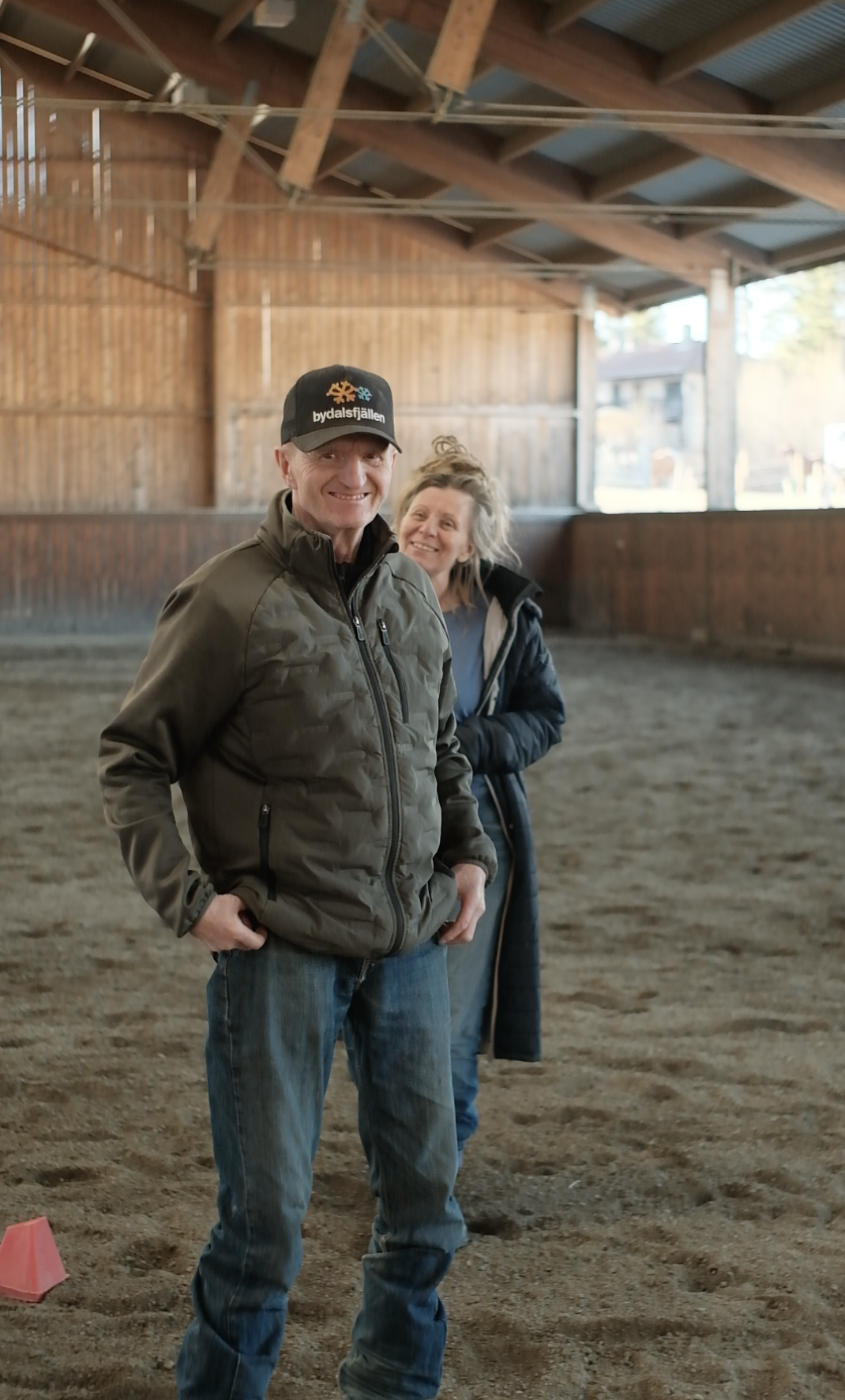 Two people standing inside a large wooden building with a dirt floor and visible roof beams, smiling and looking at the camera, during daytime.