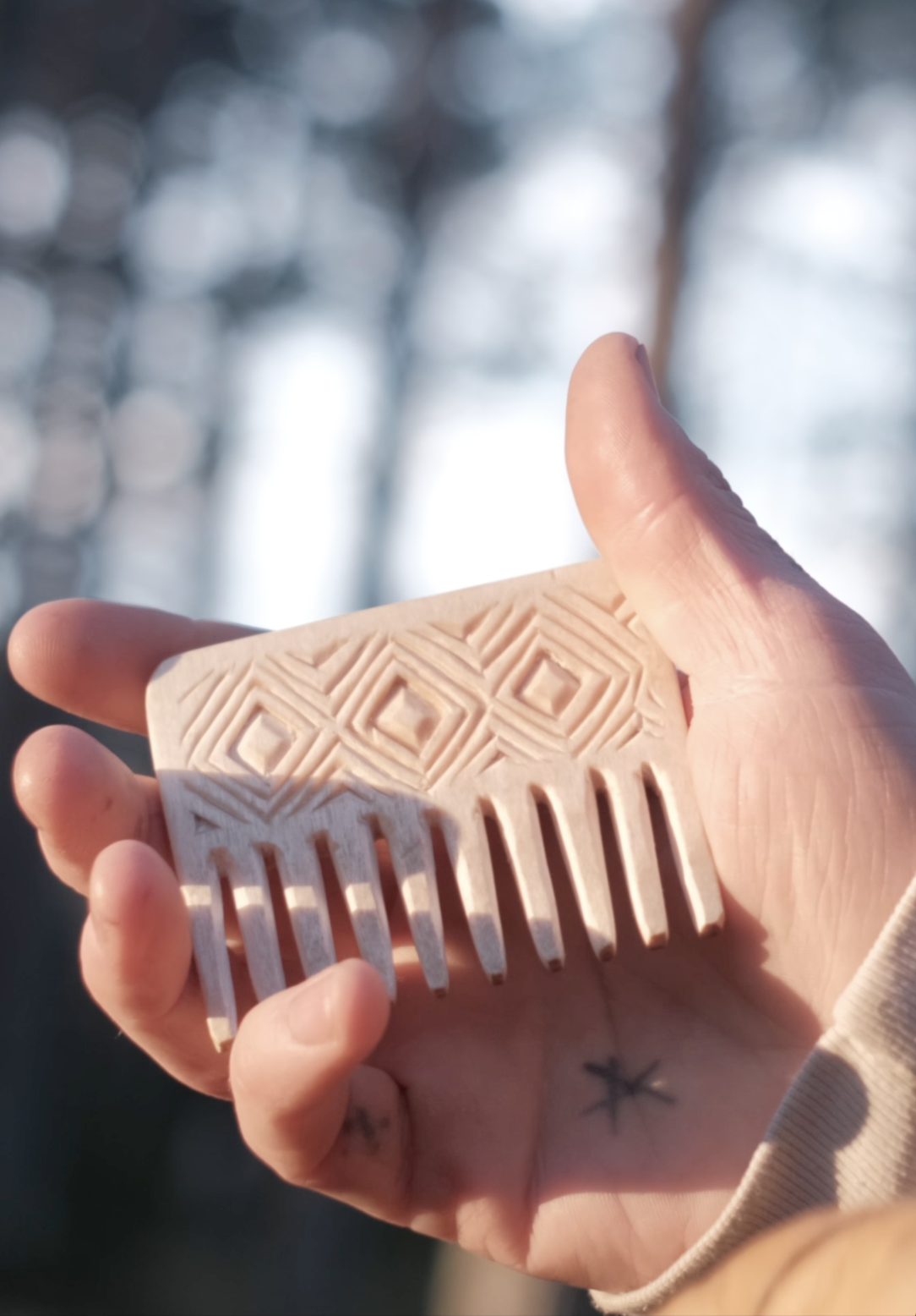 Close-up of a person holding a wooden comb with intricate geometric carvings, with a blurred outdoor background.