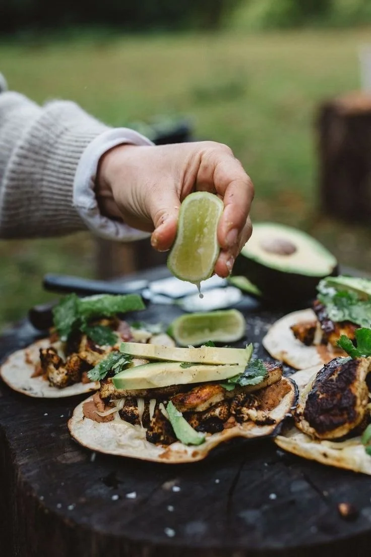 A person squeezing a lime wedge over four tacos topped with grilled meat, avocado slices, and herbs, placed on a rustic wooden surface outdoors.
