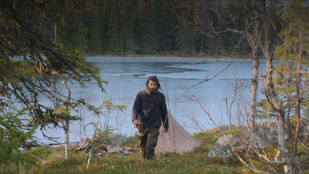 Man in dark sweater walking on grassy area near a lake with trees and tent in the background.