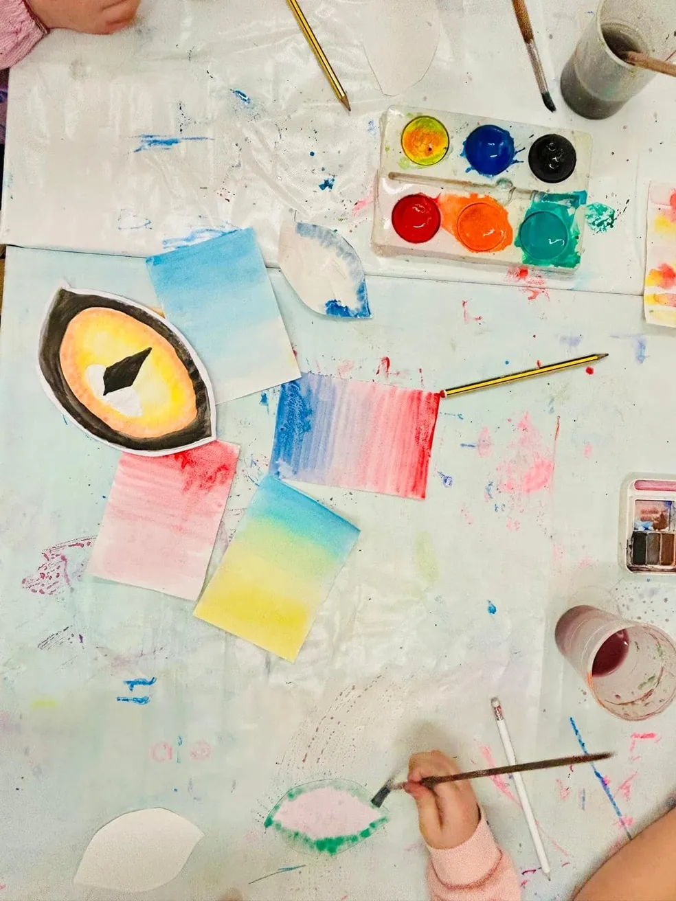 An overhead view of a table with watercolor paints, brushes, and watercolor paintings. The paintings include an eye and various colored swatches, and a child's hand is painting a green leaf shape.