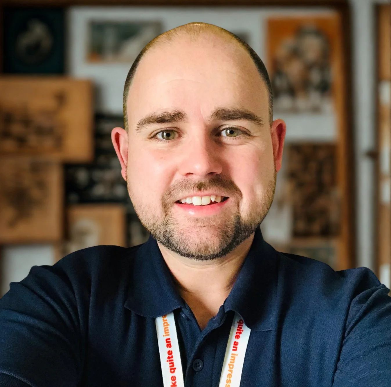 A man with a beard and receding hairline smiling at the camera, wearing a dark shirt and a lanyard around his neck, with a blurred background of framed pictures and wooden shelves.