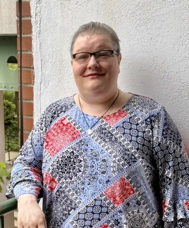 A woman with glasses and gray hair standing outside in front of a white wall, wearing a patterned dress and a necklace.