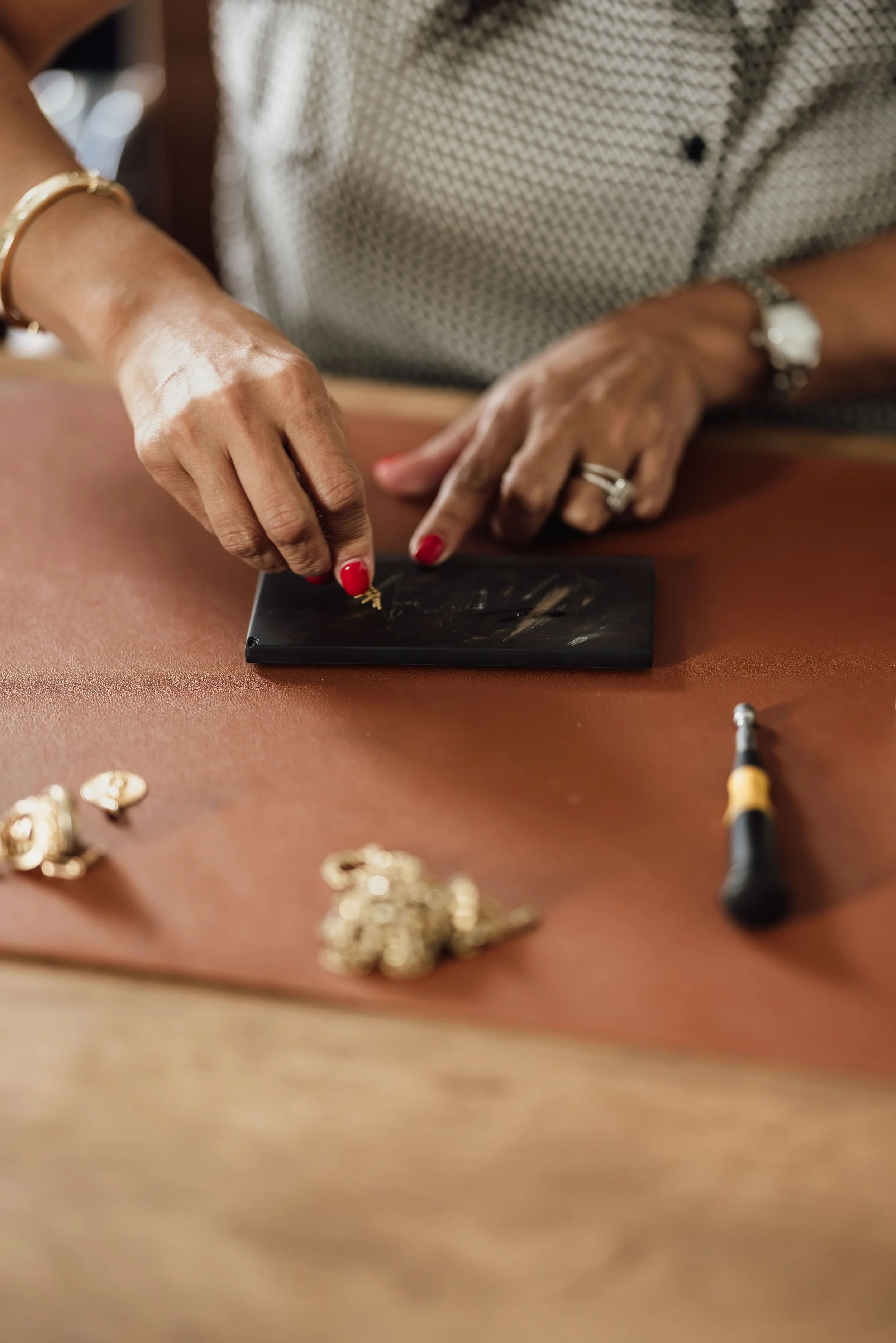 Close-up of a woman's hands working with jewelry on a workspace, with various jewelry pieces scattered around, wearing a white patterned shirt and jewelry including rings, a watch, and bracelets, with tools such as a screwdriver in the scene.