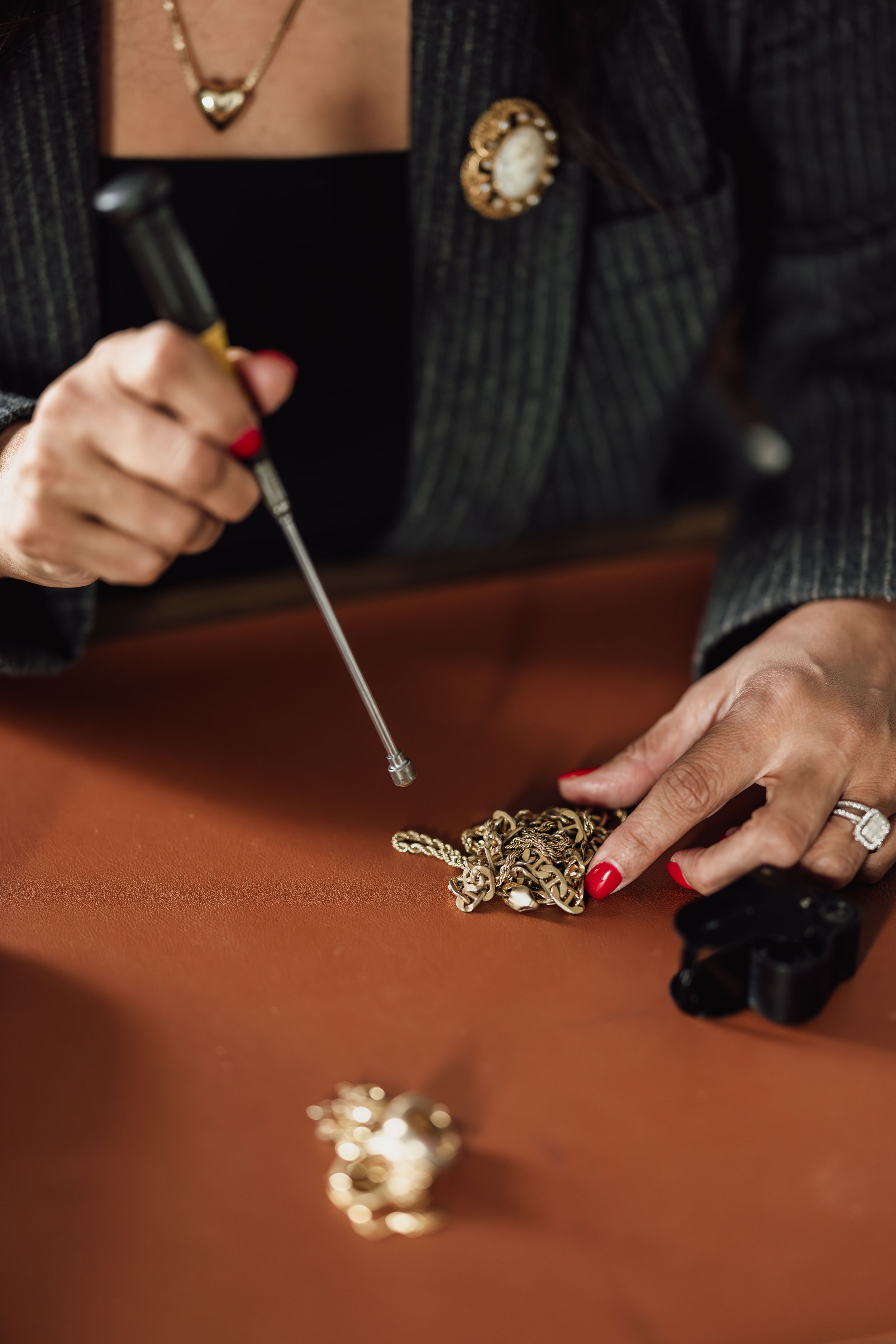 Close-up of a woman wearing a dark blazer with a decorative brooch, jewelry, and red nail polish, sorting through jewelry on a reddish-brown surface, with a jewelry screwdriver in hand.