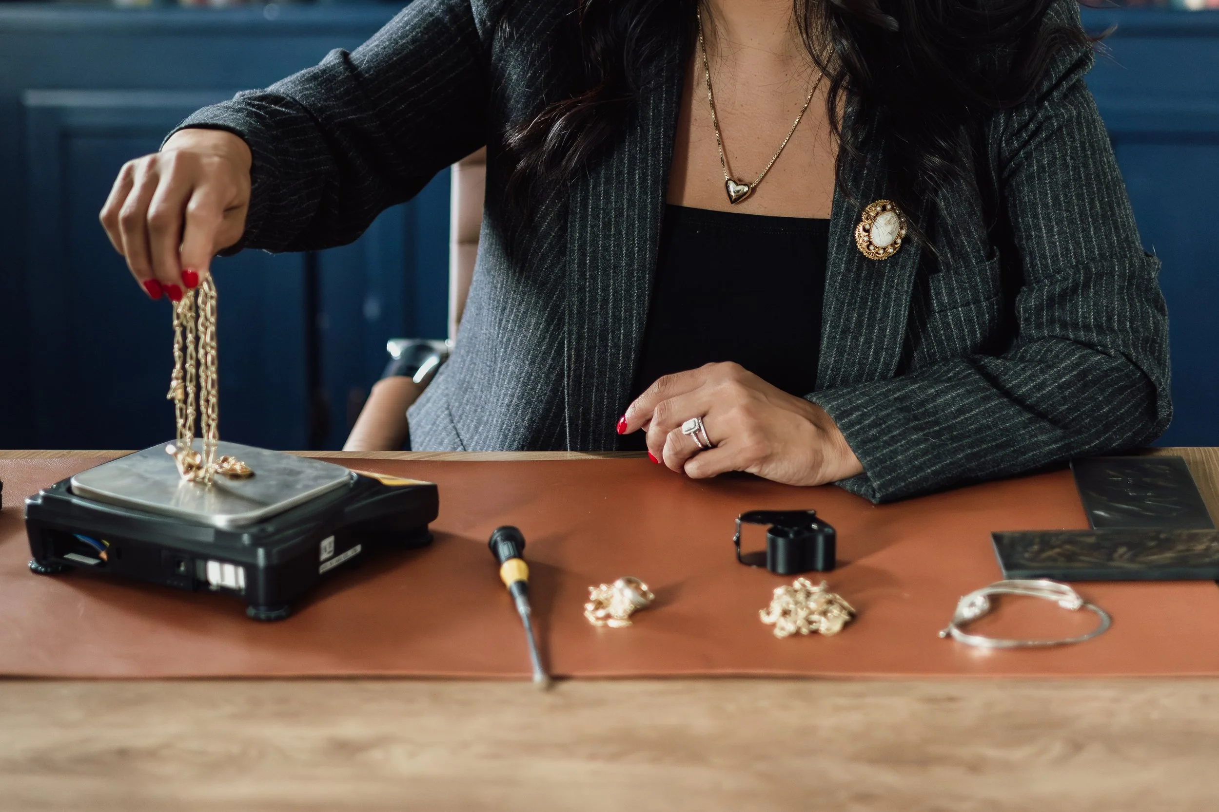 A woman wearing a dark pinstripe blazer and jewelry is sitting at a table, handling gold jewelry such as necklaces and rings, on a brown surface, with a blue background.