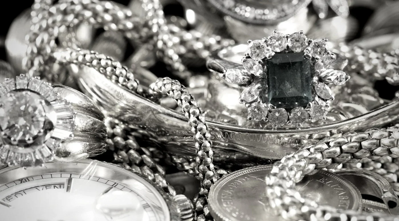 Close-up of various silver jewelry items including rings, necklaces, and a watch, with a central ring featuring a dark rectangular gemstone surrounded by small clear stones.