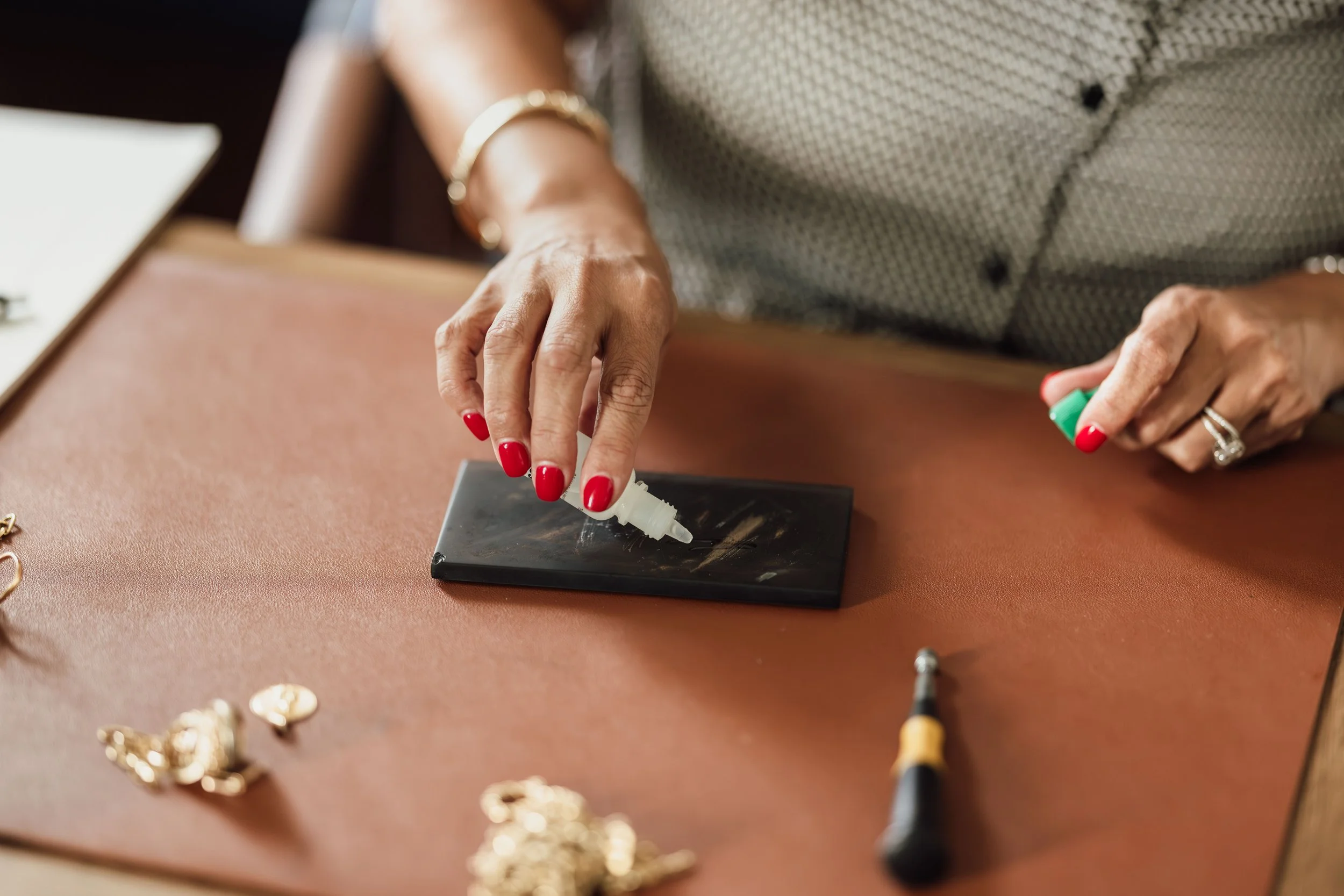 A person with red nail polish is using a small tool and a plastic syringe on a black surface, surrounded by jewelry and jewelry boxes on a wooden table.