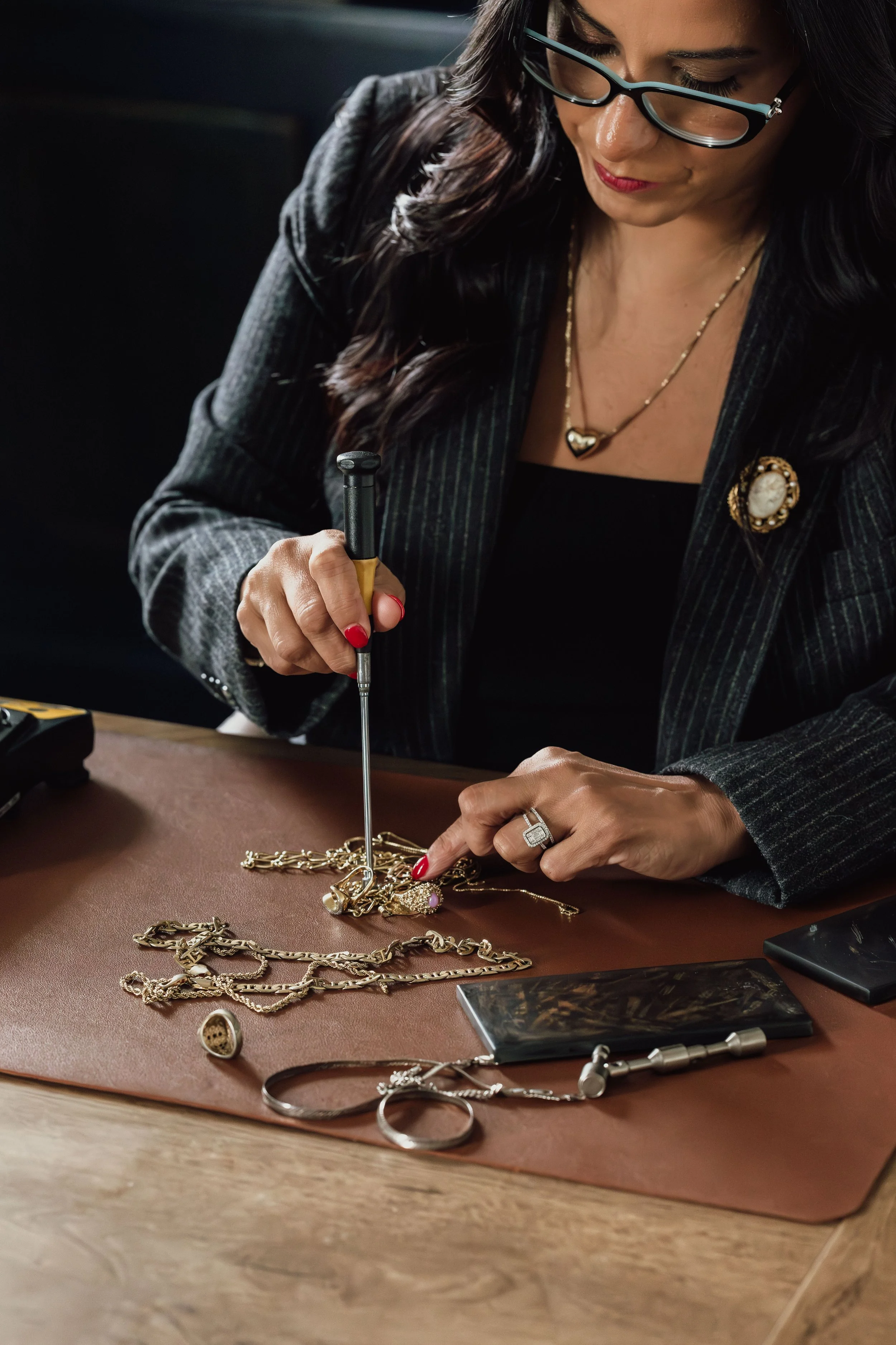 Woman with dark hair and glasses, wearing jewelry, is using a jeweler's hammer to examine or work on gold jewelry pieces and necklaces on a brown surface.