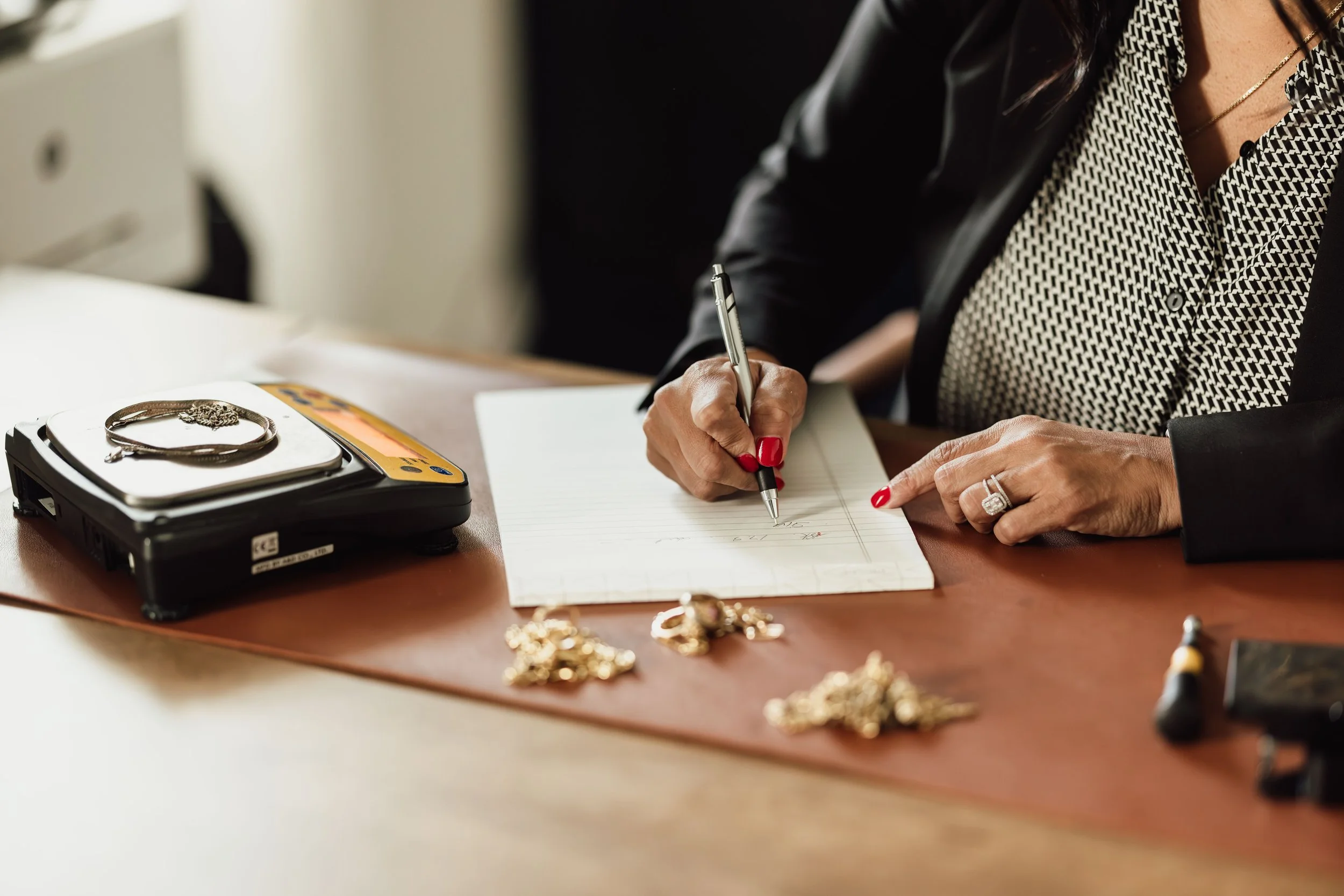 A woman wearing a black jacket and a patterned shirt is writing in a notebook at a desk. Jewelry including a bracelet and rings are on the desk, along with gold jewelry scattered on the surface and a jewelry scale with a piece of jewelry on it.