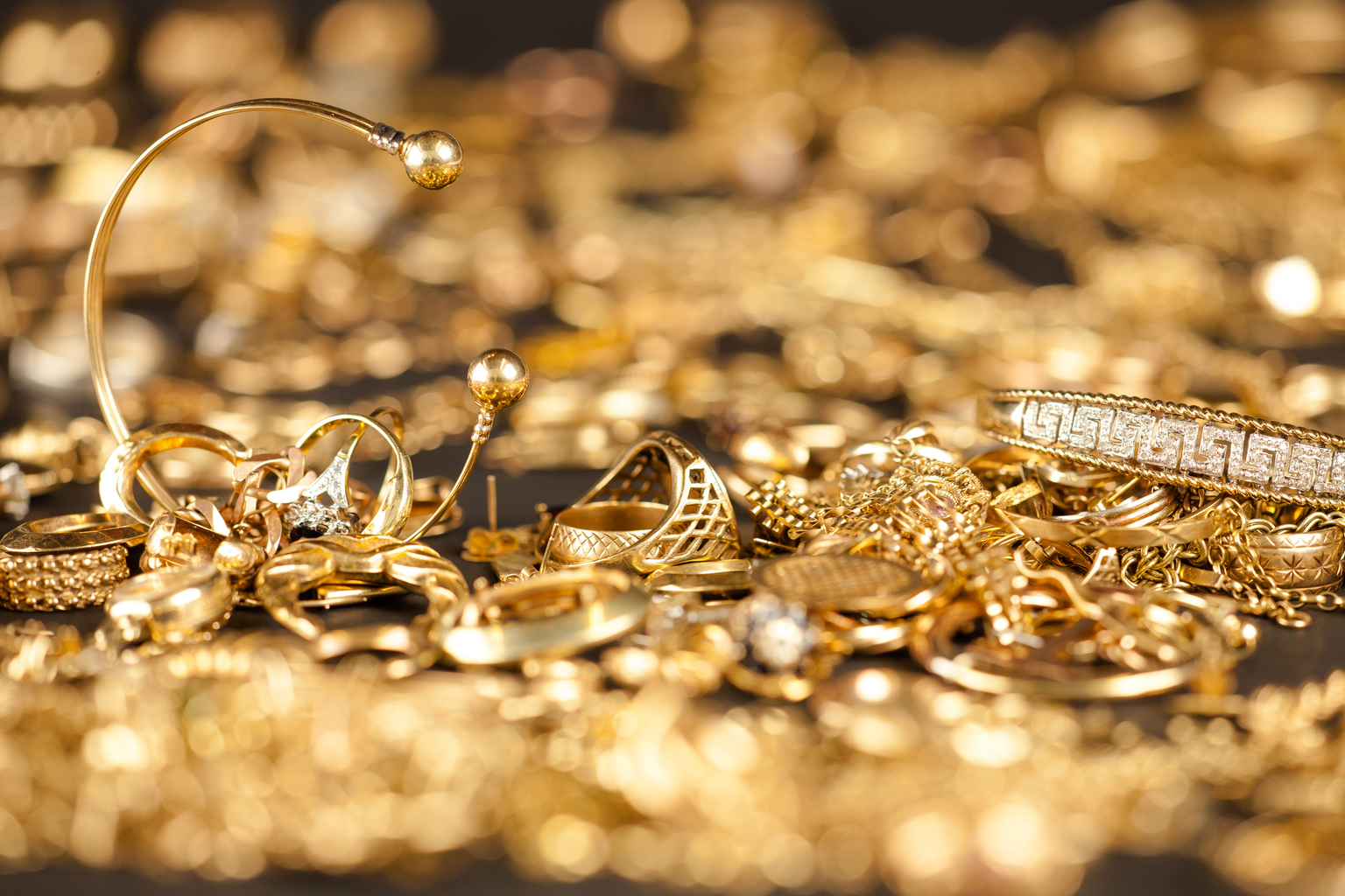 A close-up of a pile of gold jewelry including rings, bracelets, and necklaces, with a blurred background of more jewelry.