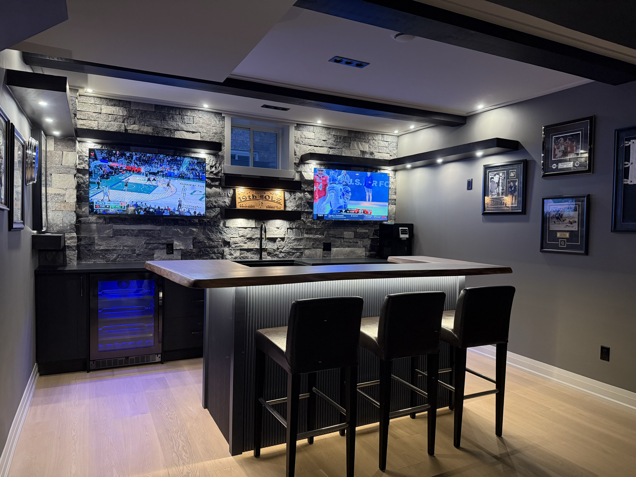 Modern basement entertainment room with two mounted flat-screen TVs showing basketball games, gray stone accent wall, black bar with high stools, wine cooler, framed sports memorabilia, and hardwood flooring.