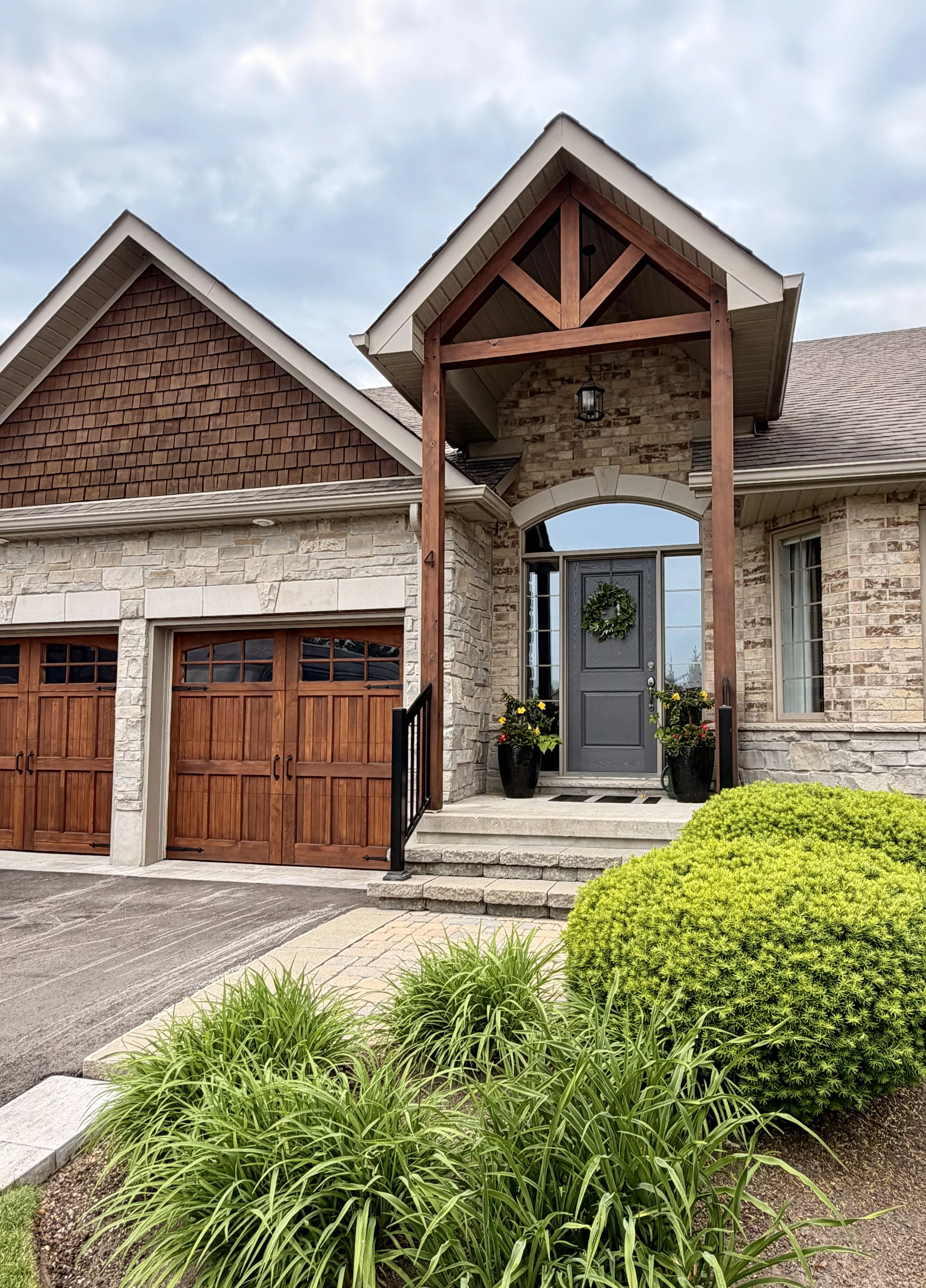 Front view of a house with a stone and brick facade, wooden garage doors, a covered porch with a gabled roof, and a gray front door decorated with a wreath. There are potted plants at the entrance, stairs leading up to the door, and landscaping with bushes and ornamental grass in the foreground.