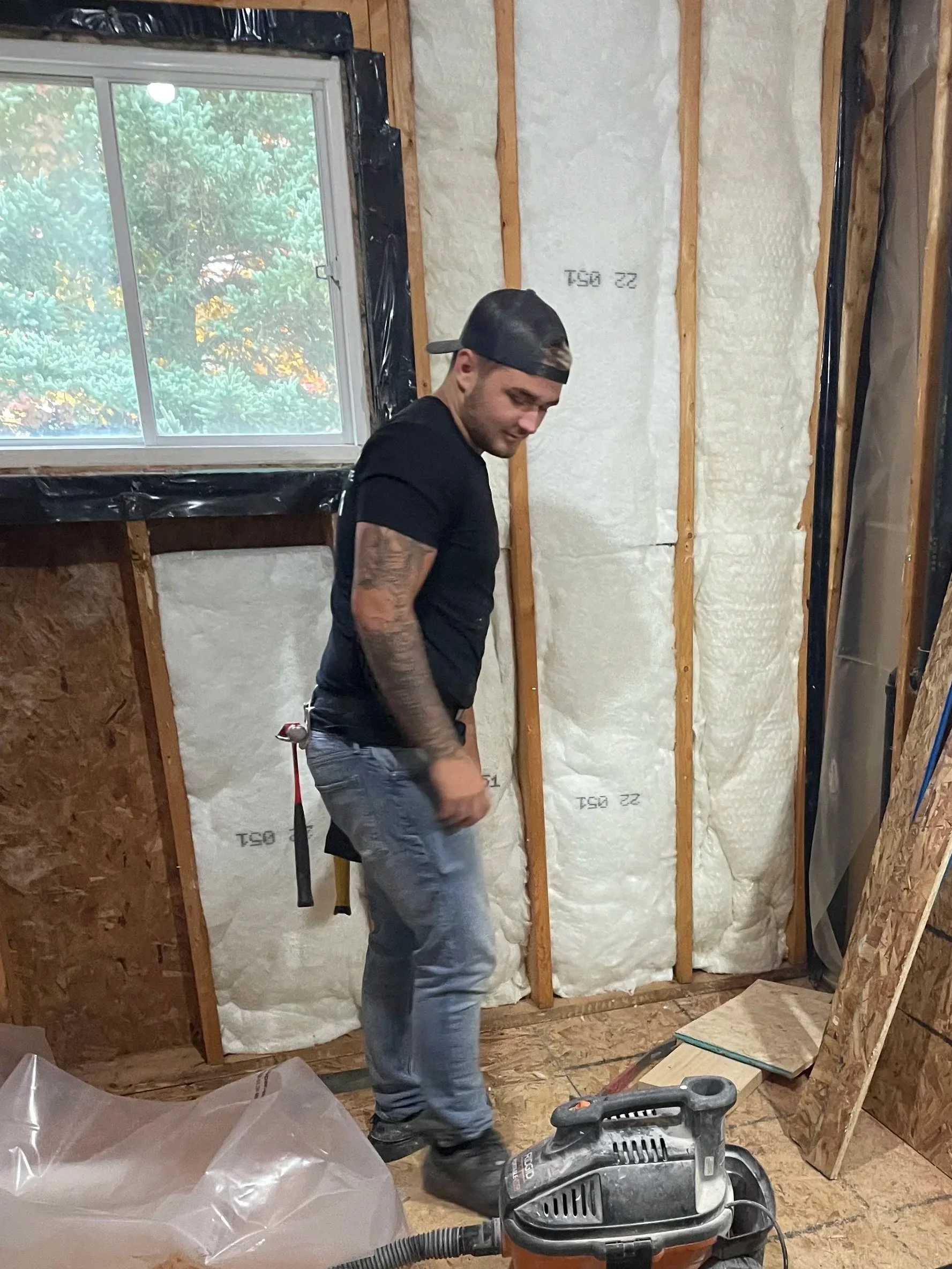 A man in a black t-shirt, jeans, and a backwards cap is working inside a room under construction, with insulation and wooden framing visible on the walls, and construction tools on the floor.