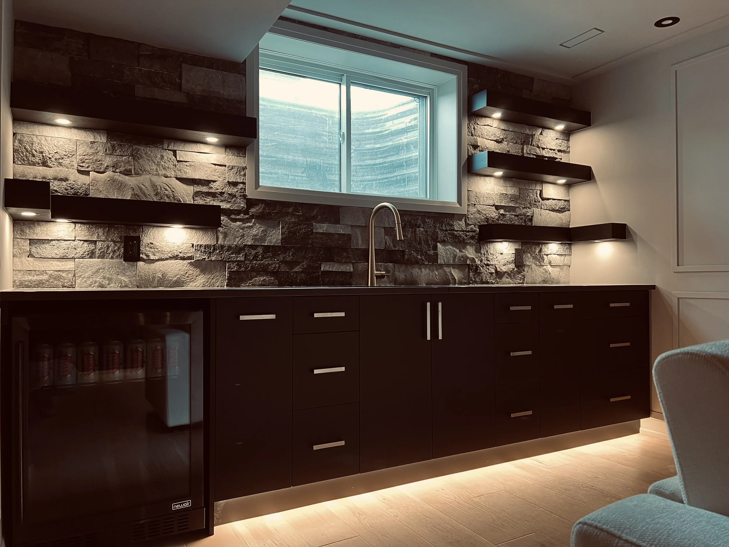 Modern kitchen with stone accent wall, white window, dark cabinets, built-in lighting, and a mini fridge.