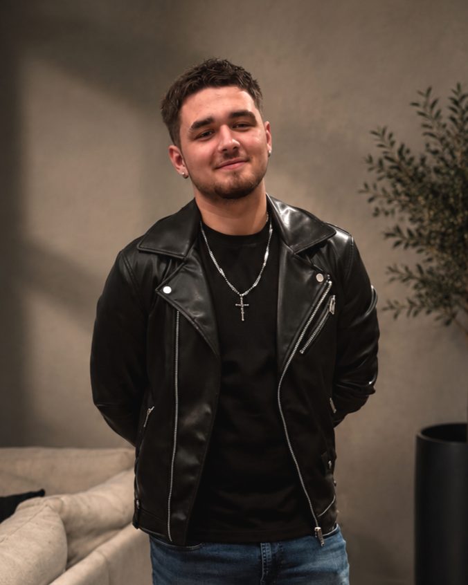 A young man with short brown hair and a light beard, wearing a black leather jacket, black t-shirt, jeans, silver chain with a cross pendant, and earrings, standing indoors with a sofa and a potted plant in the background.