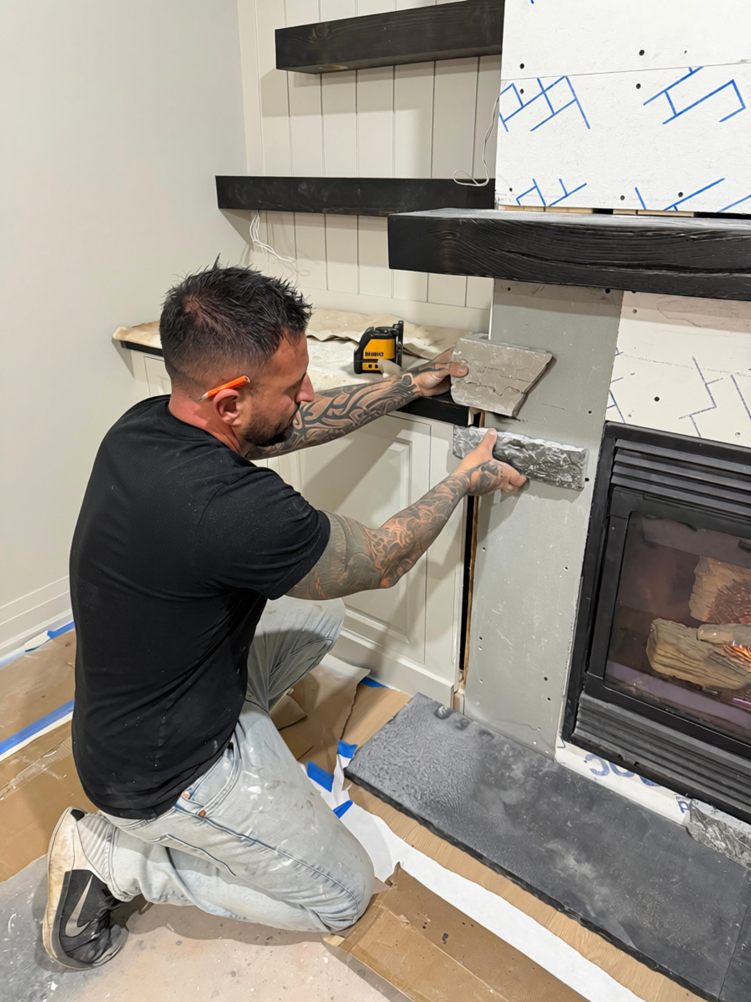A man with tattoos on his arms installing a fireplace or stove in a house under construction. He is kneeling and holding tiles, with a toolbox and construction materials around.